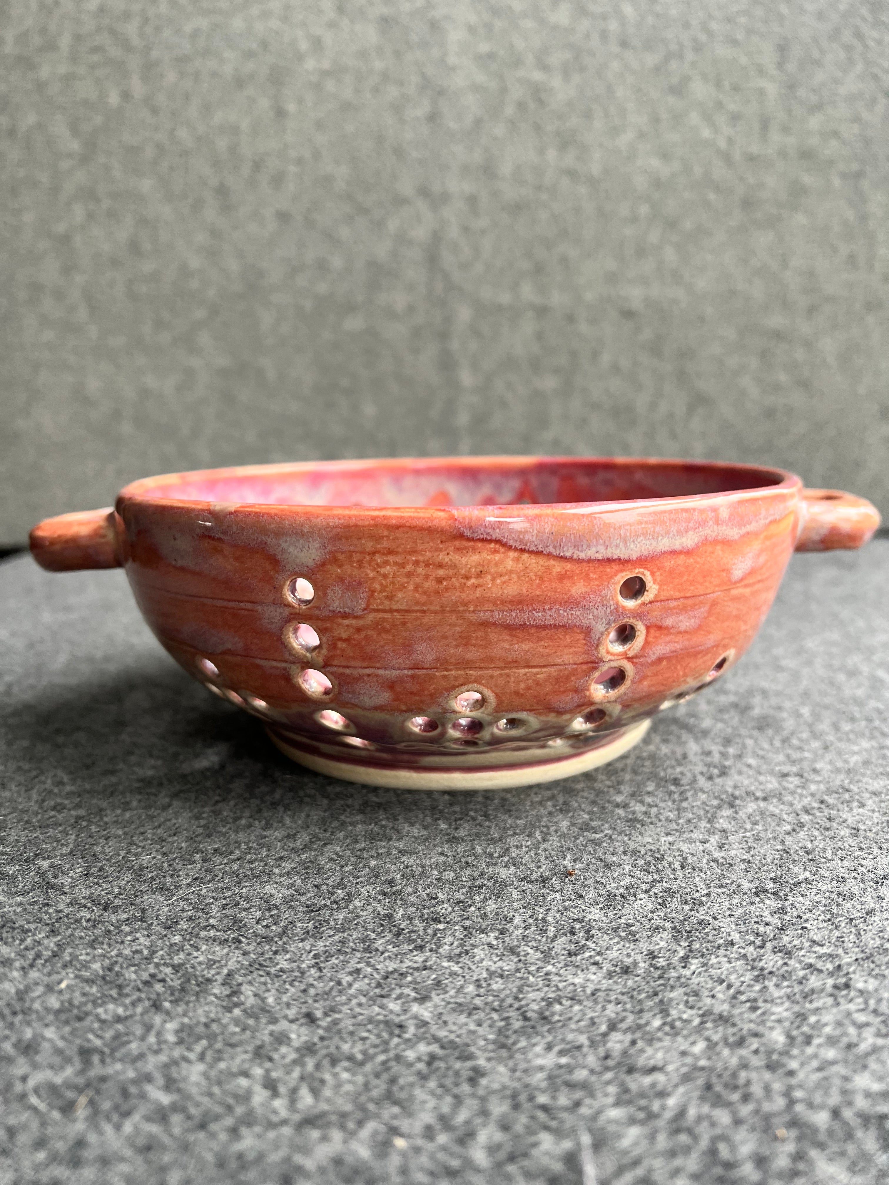 A side view of a pink bowl with two small loop handles, and intricate pierced holes and a bright red heart at the base sits in front of a grey background.