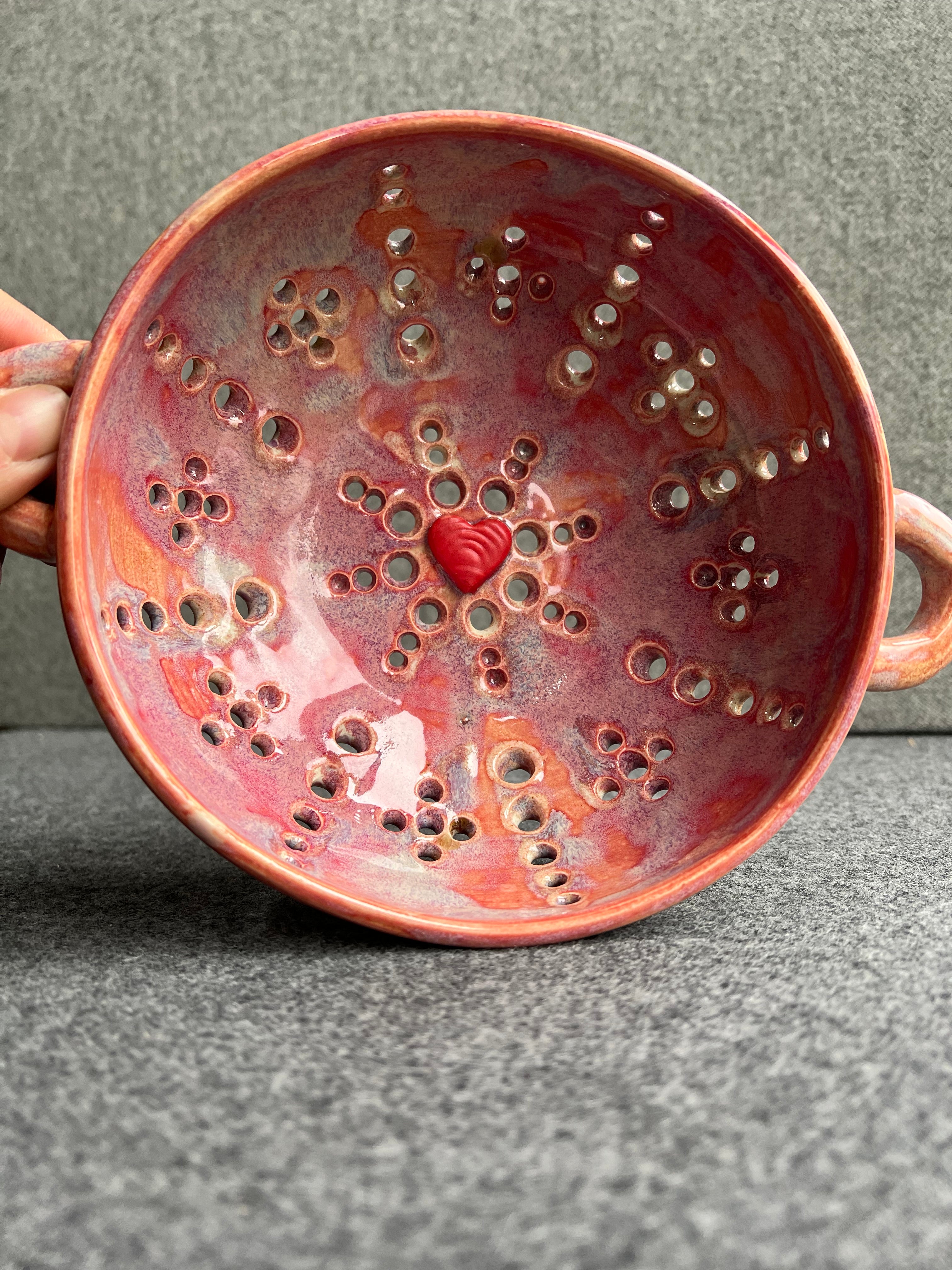 A pink bowl with two small loop handles, and intricate pierced holes and a bright red heart at the base sits in front of a grey background.