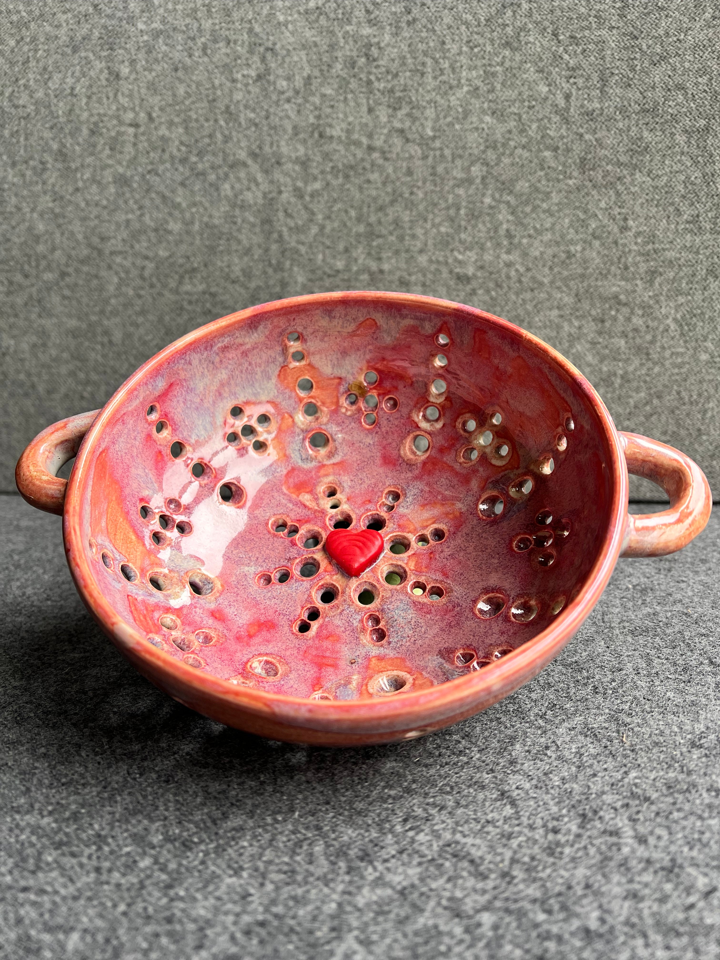 A pink bowl with two small loop handles, and intricate pierced holes and a bright red heart at the base sits in front of a grey background.