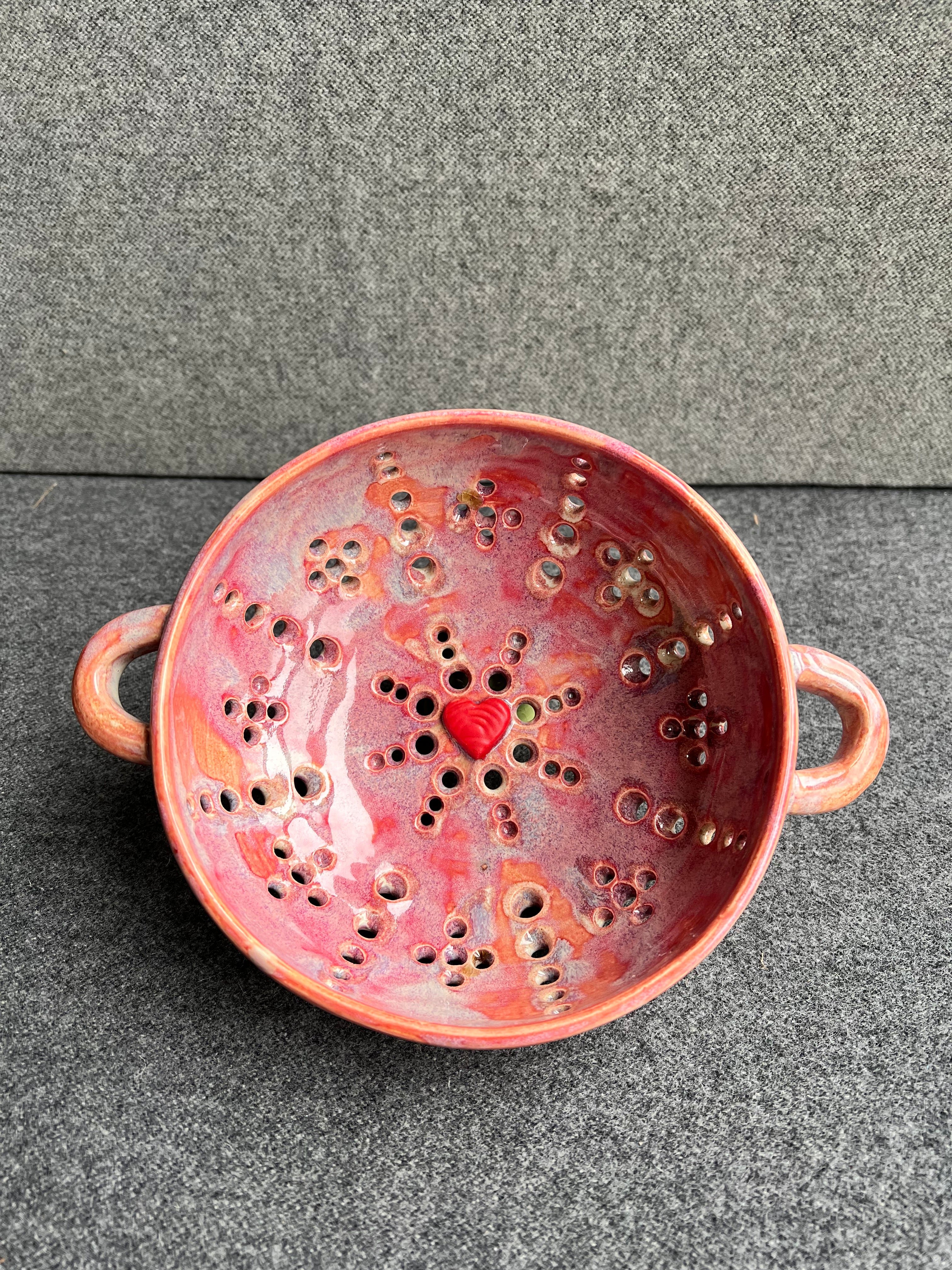 A pink bowl with two small loop handles, and intricate pierced holes and a bright red heart at the base sits in front of a grey background.