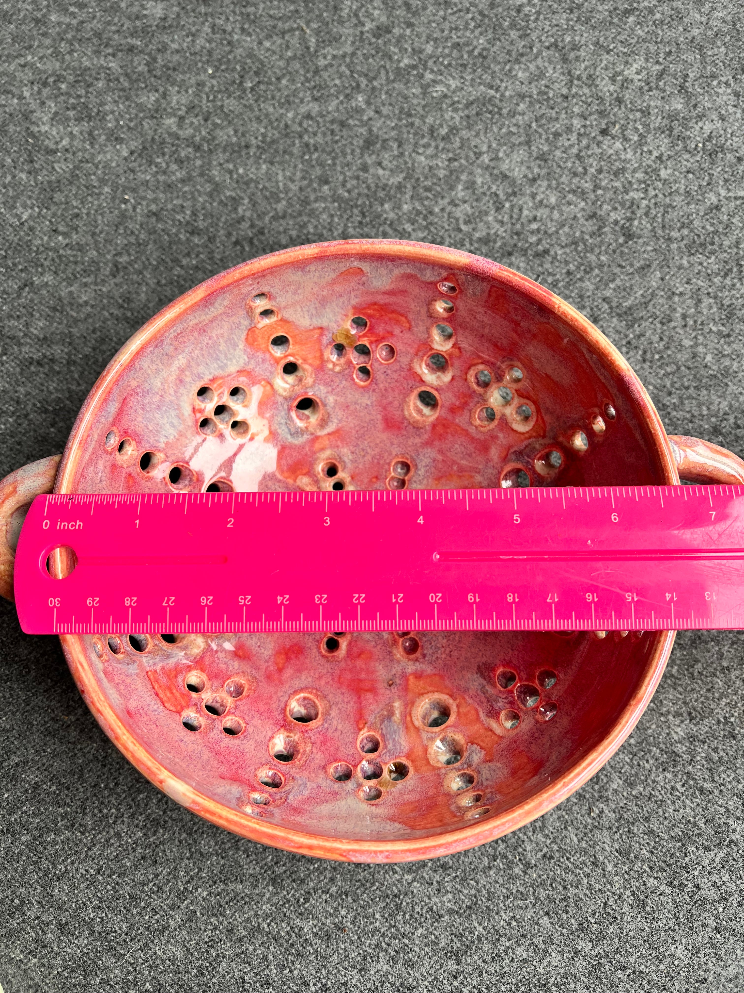 A bright pink ruler sits on top of a pink bowl with two small loop handles, and intricate pierced holes and a bright red heart at the base sits in front of a grey background. The ruler measure 6.75 inches.