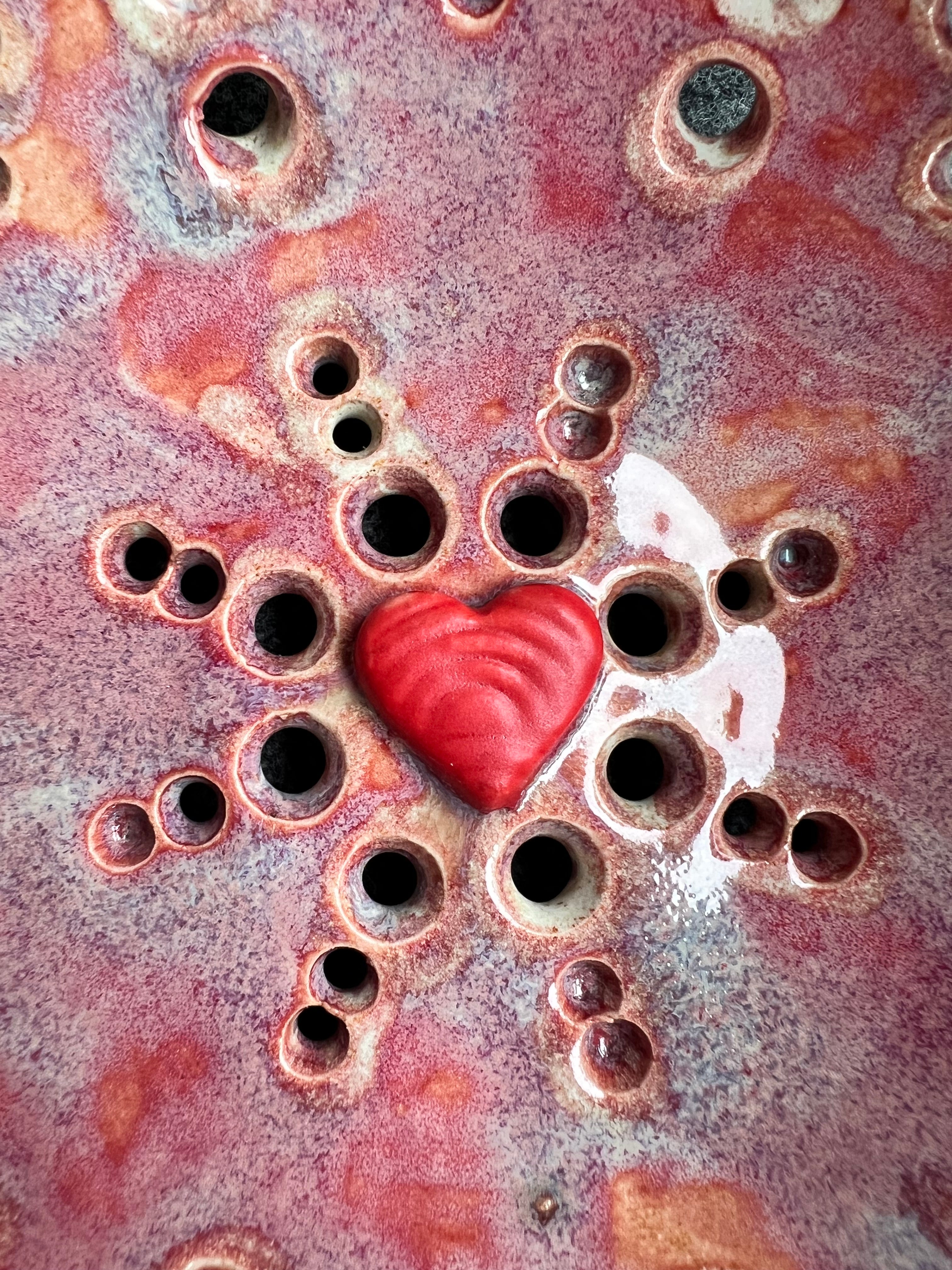 A close up of a pink bowl with two small loop handles, and intricate pierced holes and a bright red heart at the base sits in front of a grey background.
