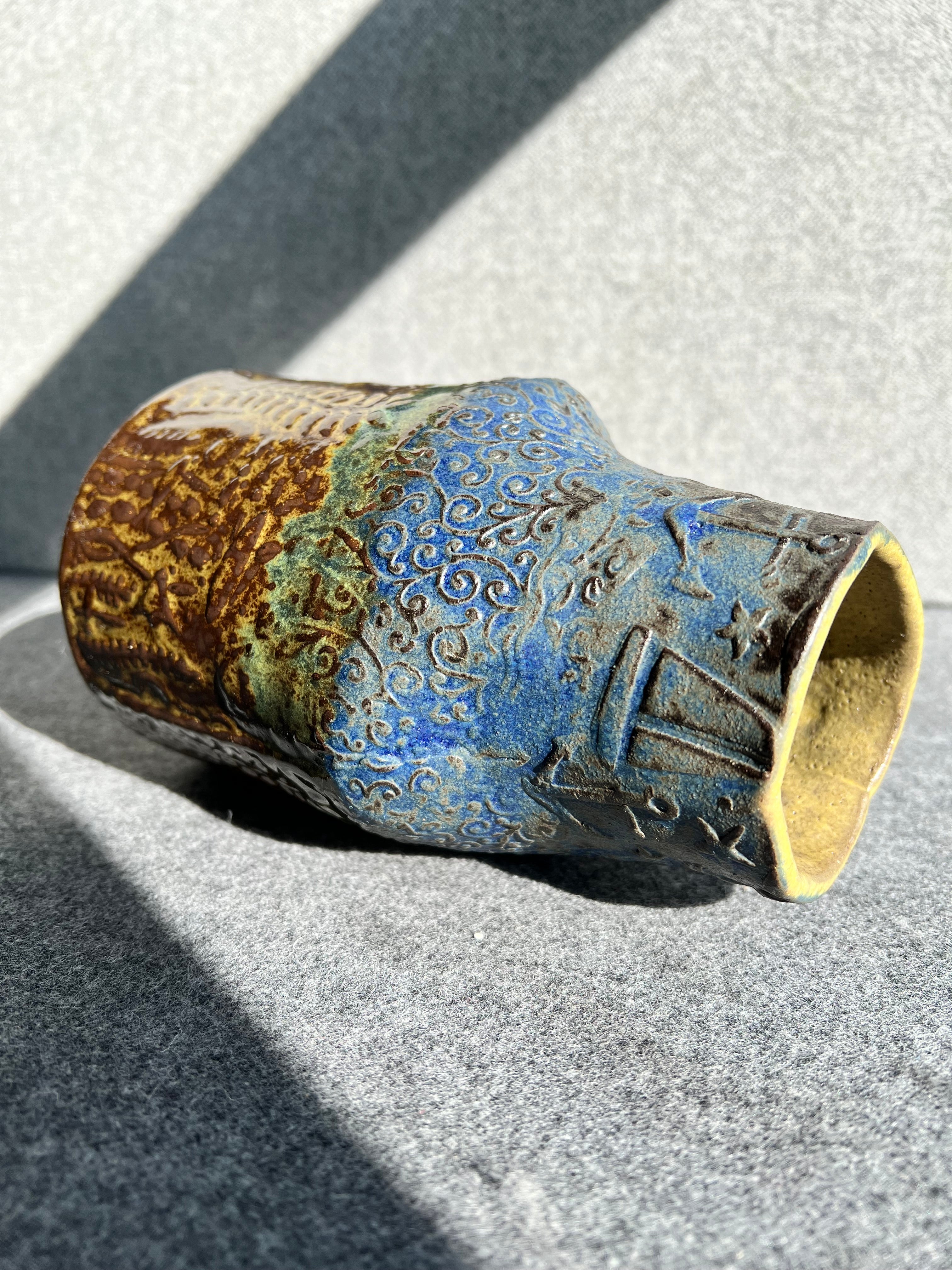 a three sided, handbill, green, brown, and blue ocean themed vase lays on its side in front of a grey background.