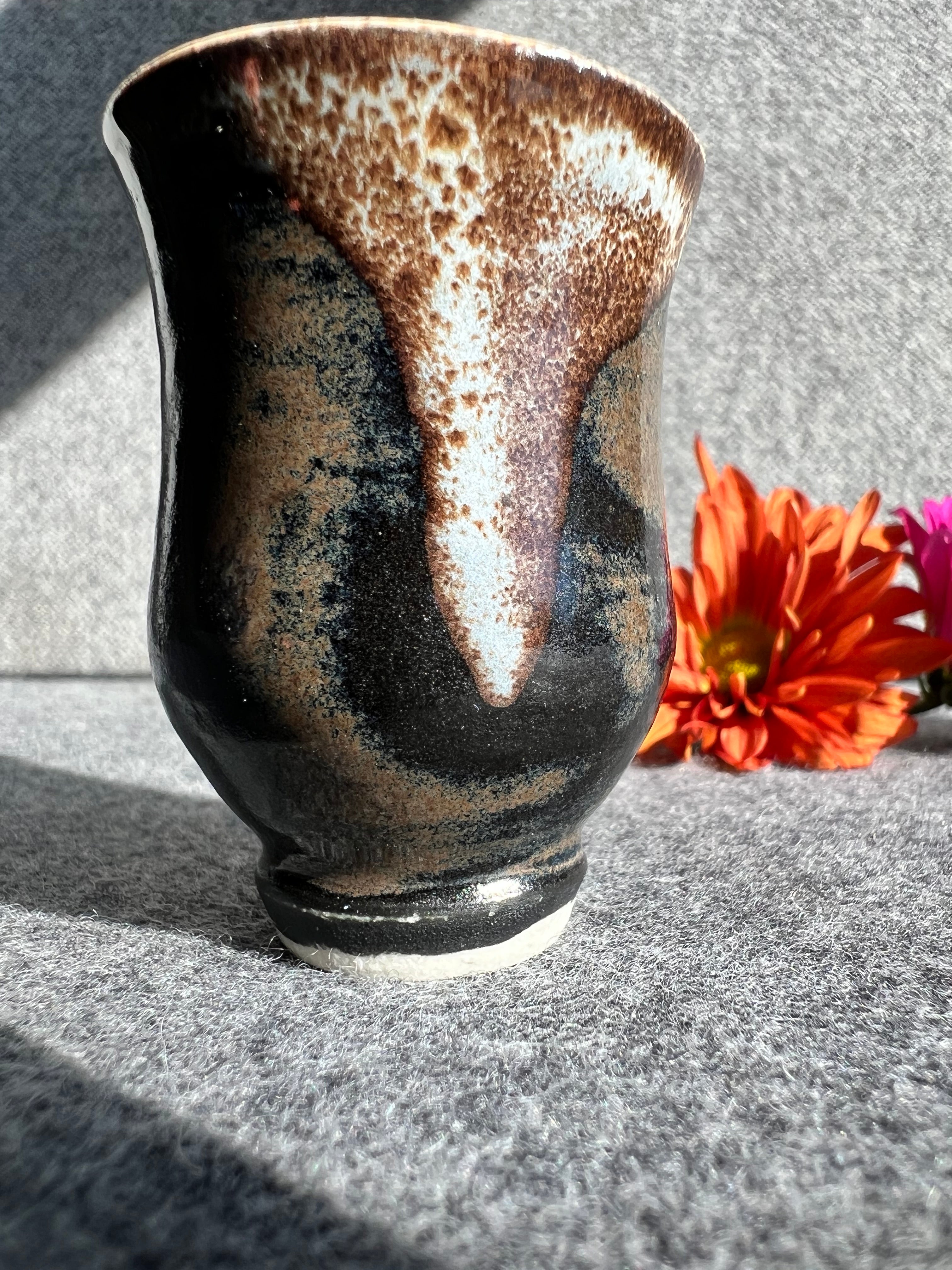 A curvaceous black, brown, and cream colored bud vase sits in front of two out of focus flowers and a grey background.
