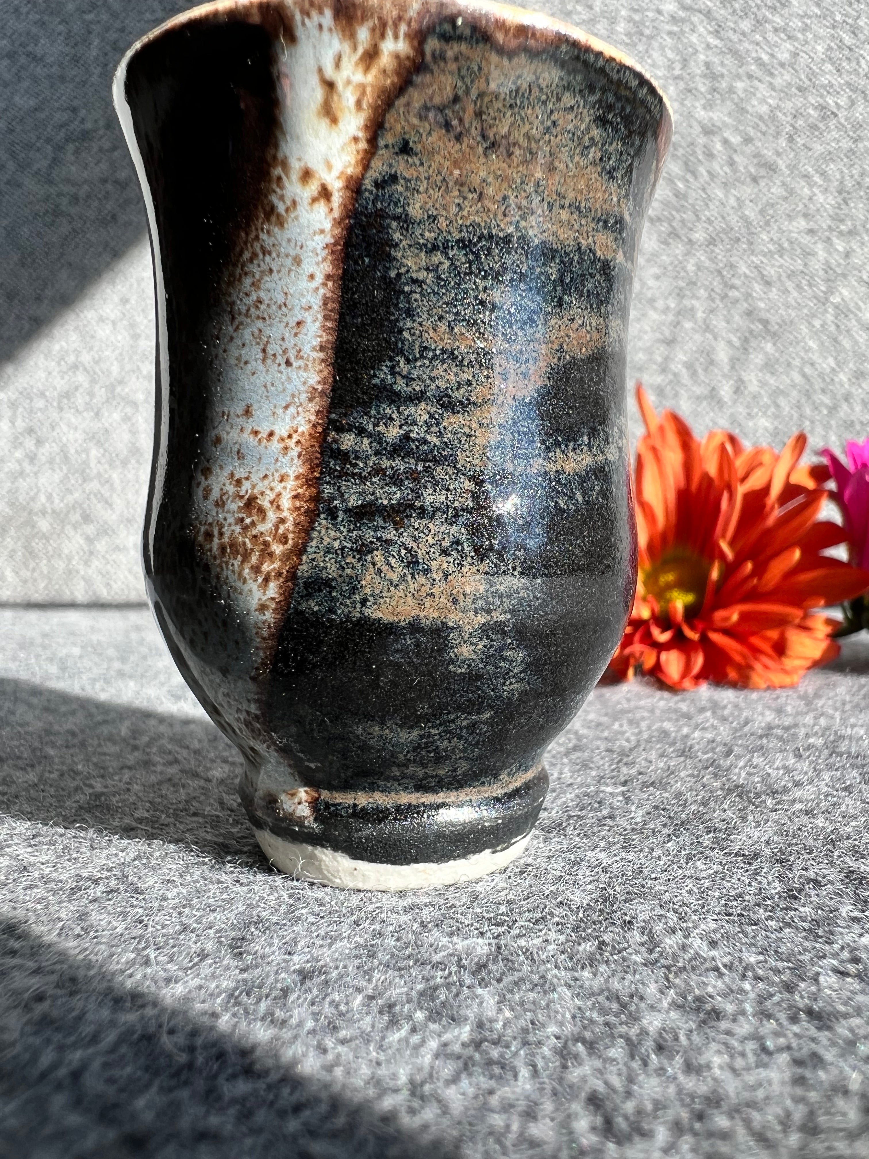 A curvaceous black, brown, and cream colored bud vase sits in front of two out of focus flowers and a grey background.