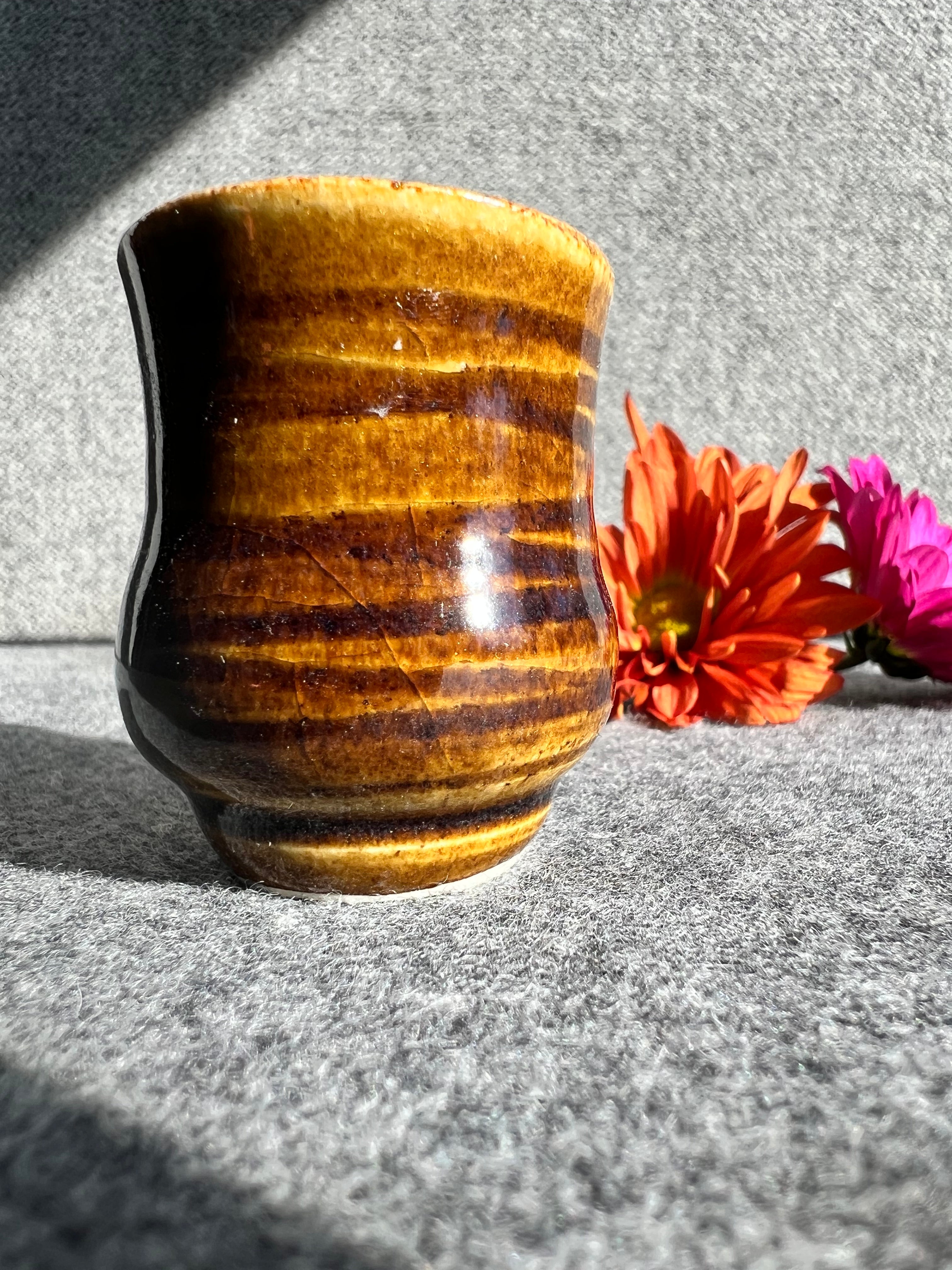 A rusty iron colored, glazed bud vase with a swirly texture sits in front of two out of focus flowers and a grey background.