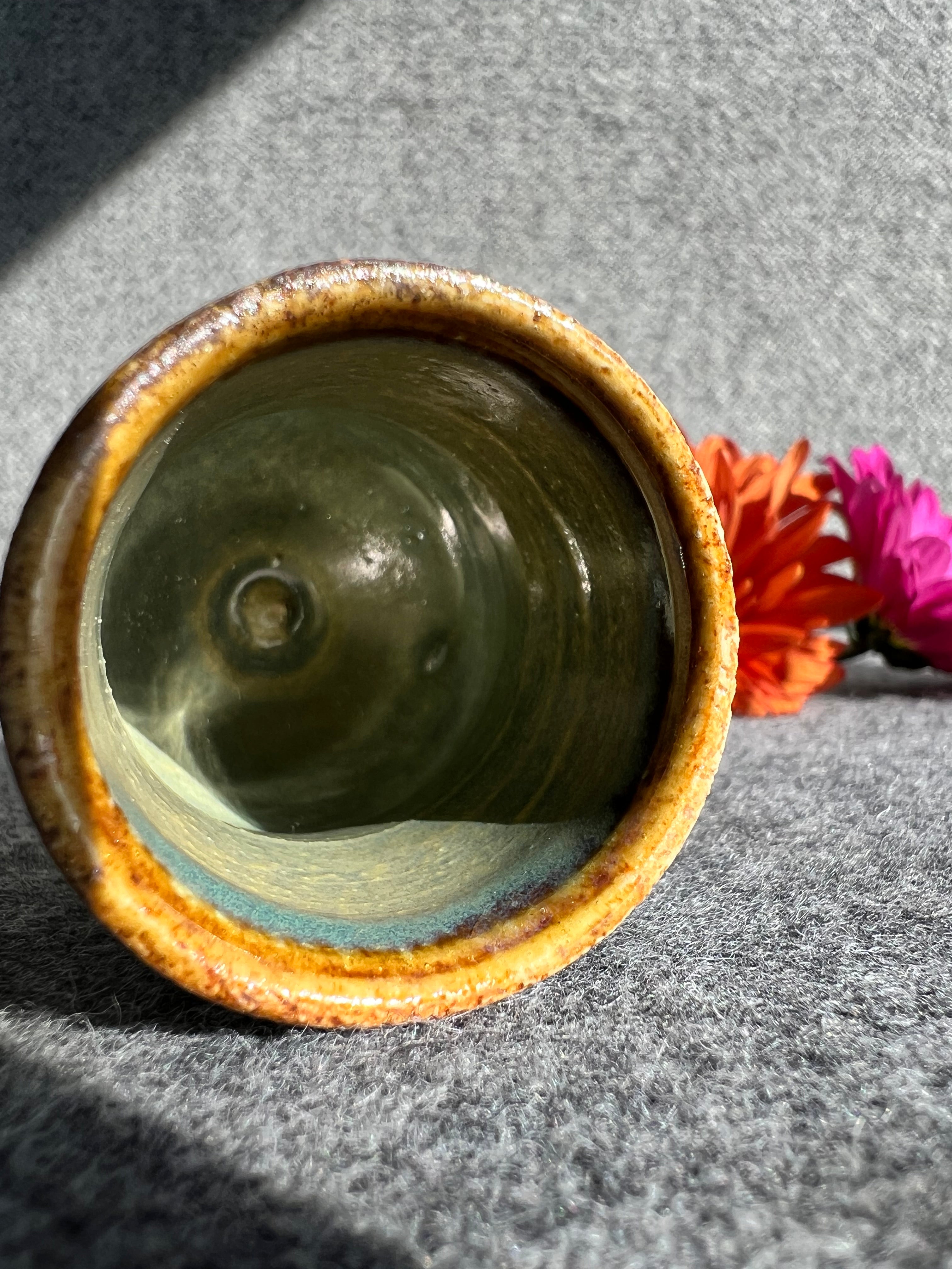 A rusty iron colored, glazed bud vase with a swirly texture lays on its side in front of two out of focus flowers and a grey background.