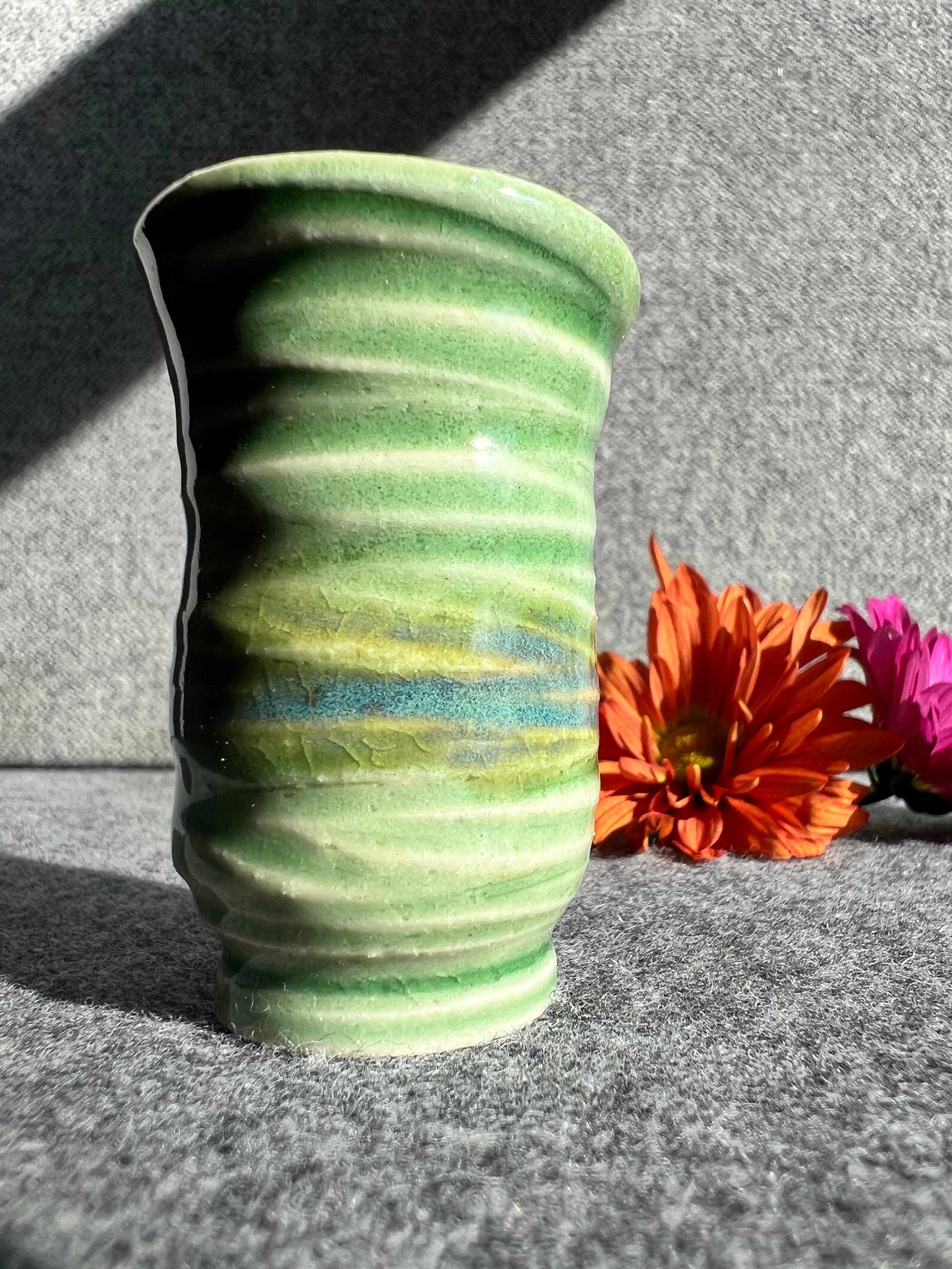 A tall thin bud vase with a swirly texture and green and blue glaze sits in front of two out of focus flowers and a grey background.