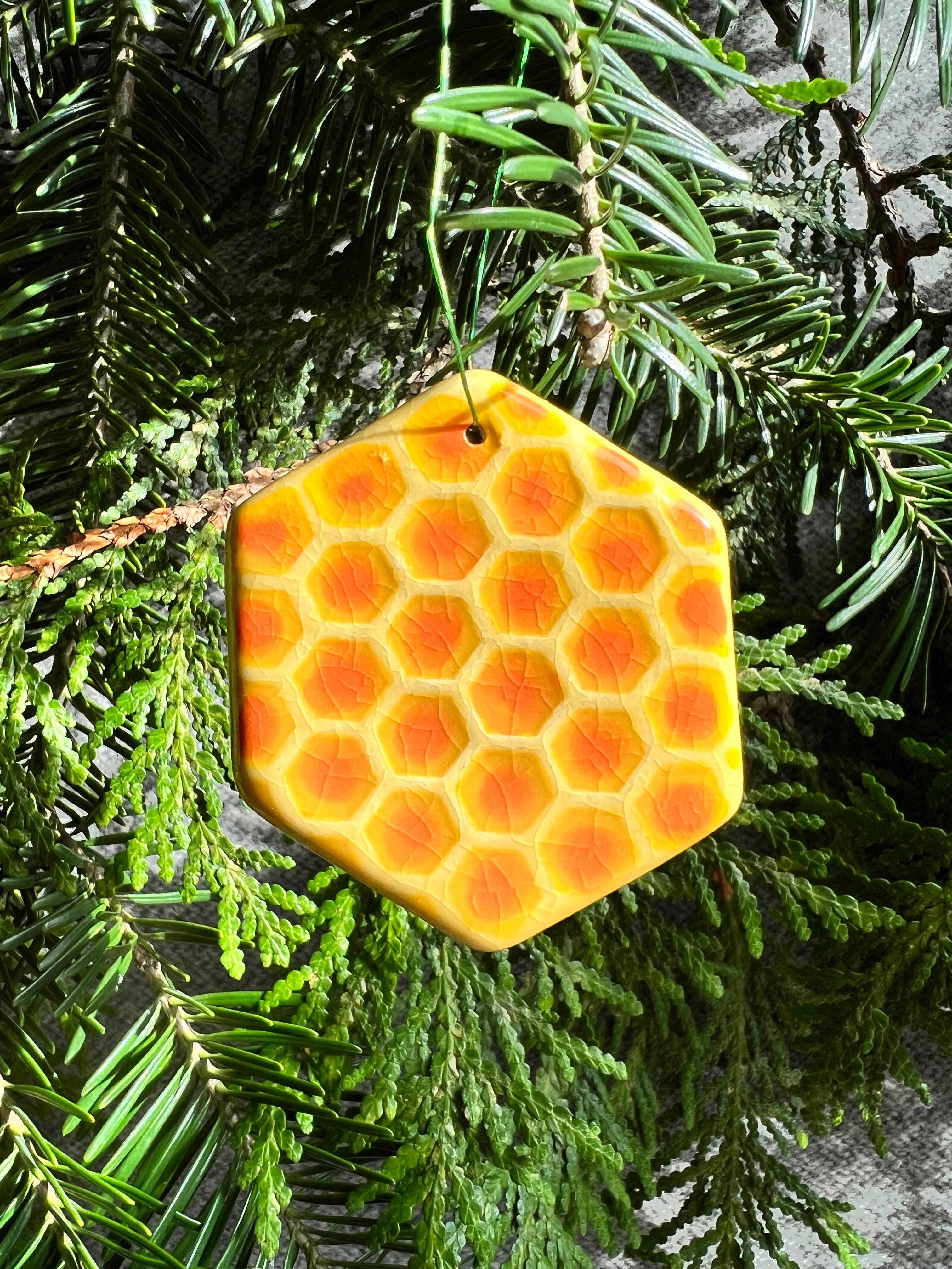 A yellow and orange hexagon with a crackle clear glaze and textured honeycomb surface ornament hangs in front of cedar and balsam branches.