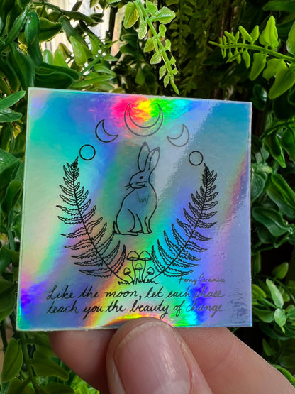 A person is holding up a Ferny Ceramics 2x2 Square Holographic Sticker with an image of a rabbit, highlighting its resilience to water damage.
