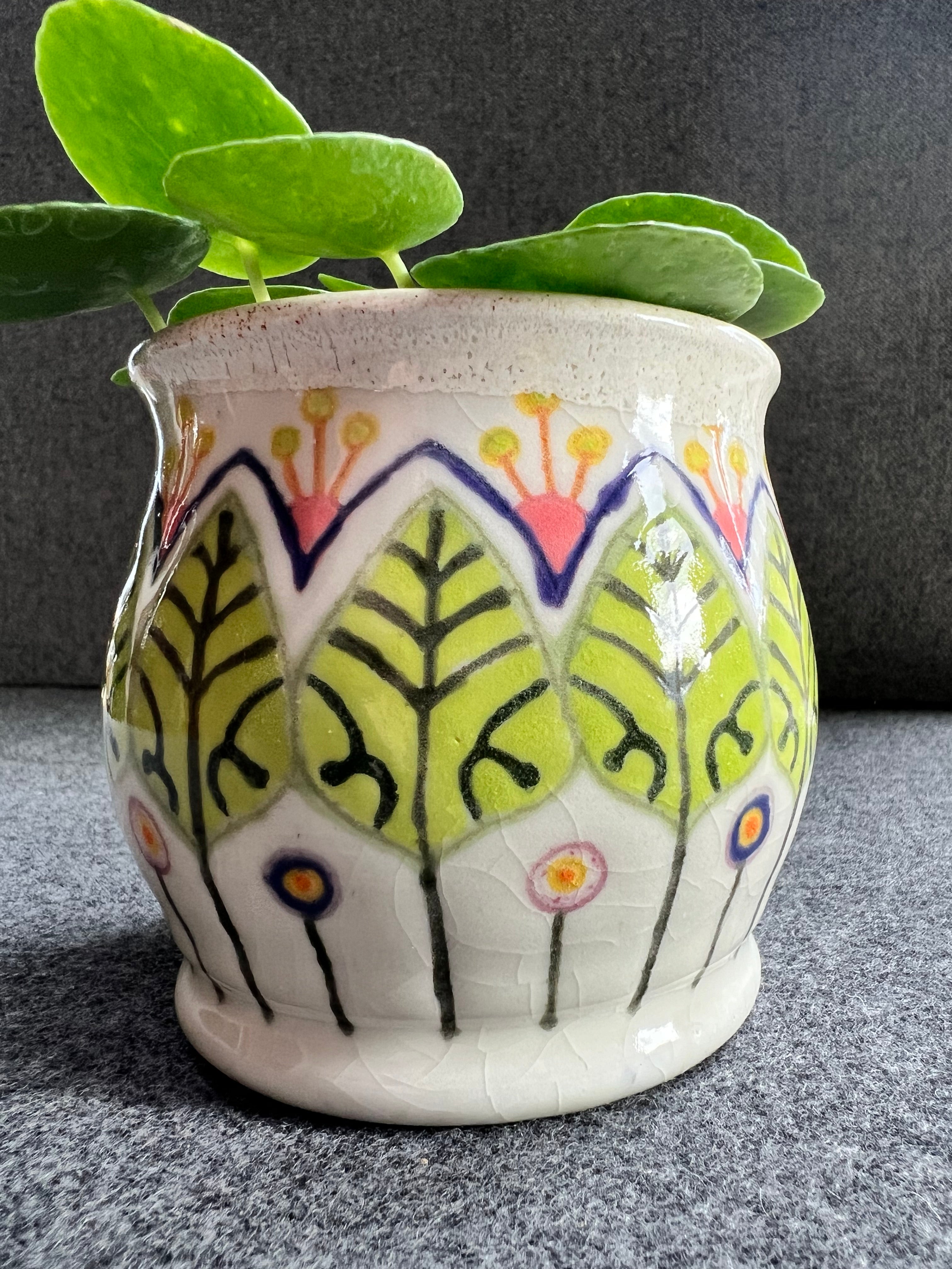 A white ceramic pot with green leaf pattern and circular abstract flowers holds a lush green plant.