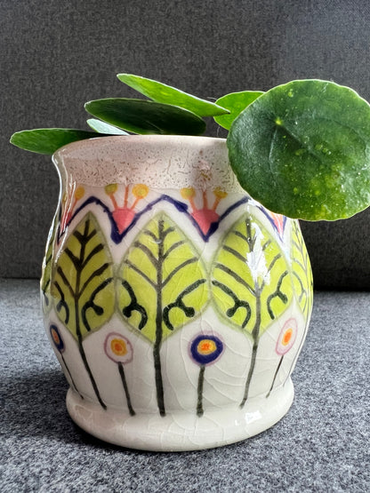 A white ceramic pot with green leaf and circular abstract flowers holds a lush green plant.