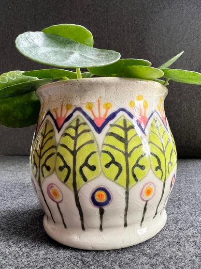 A white ceramic pot with green leaf and circular abstract flowers holds a lush green plant.