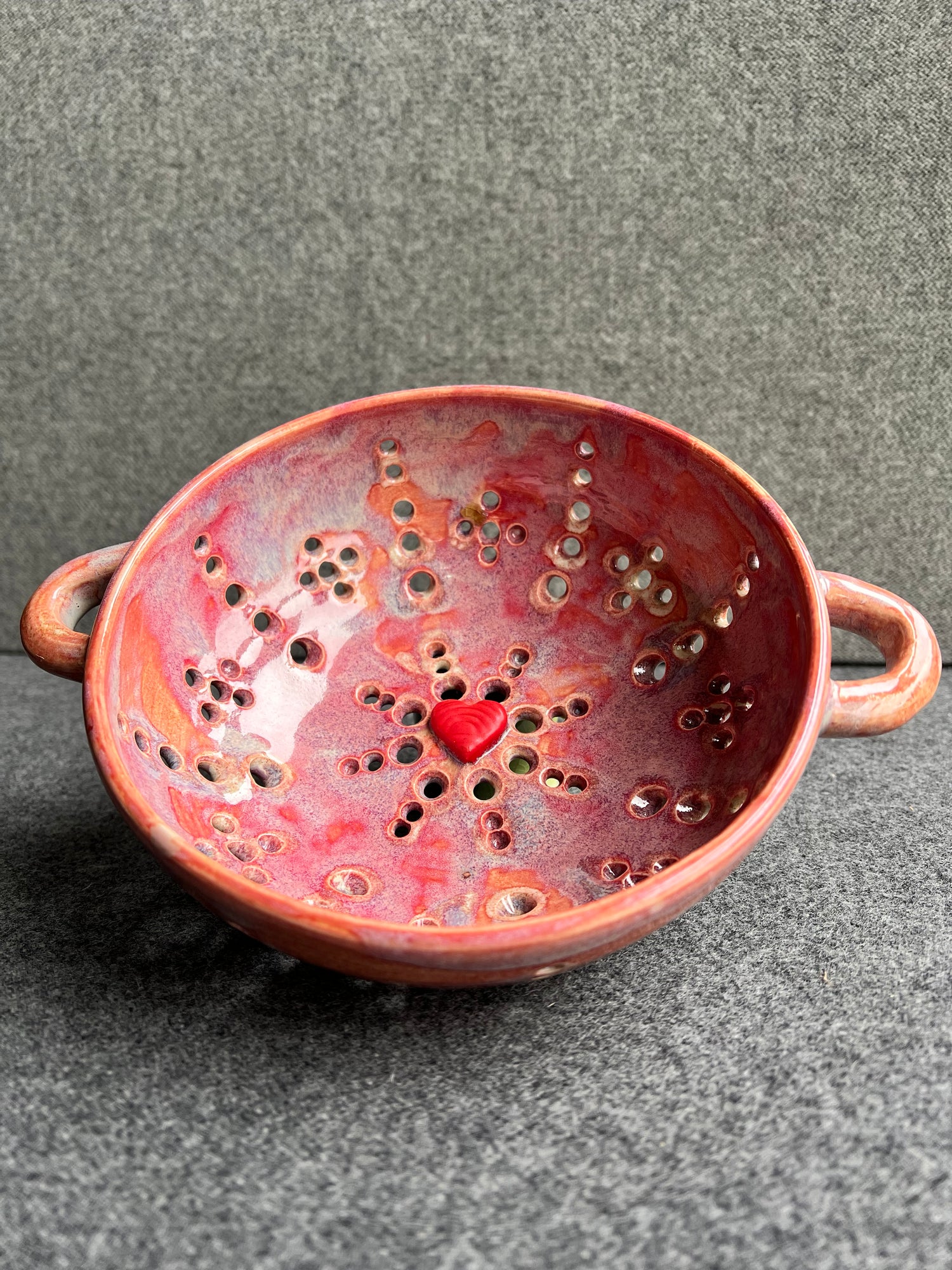 A pink bowl with two small loop handles, and intricate pierced holes and a bright red heart at the base sits in front of a grey background. 