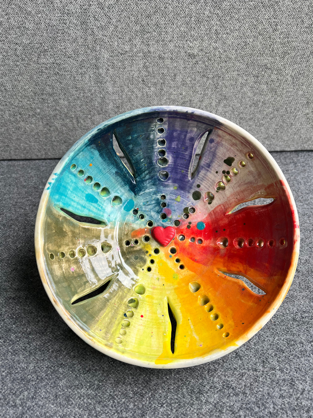 An intricately pierced bowl with a color wheel effect rainbow pattern and a small red heart in the center of the bowl sits in front of a grey background. 