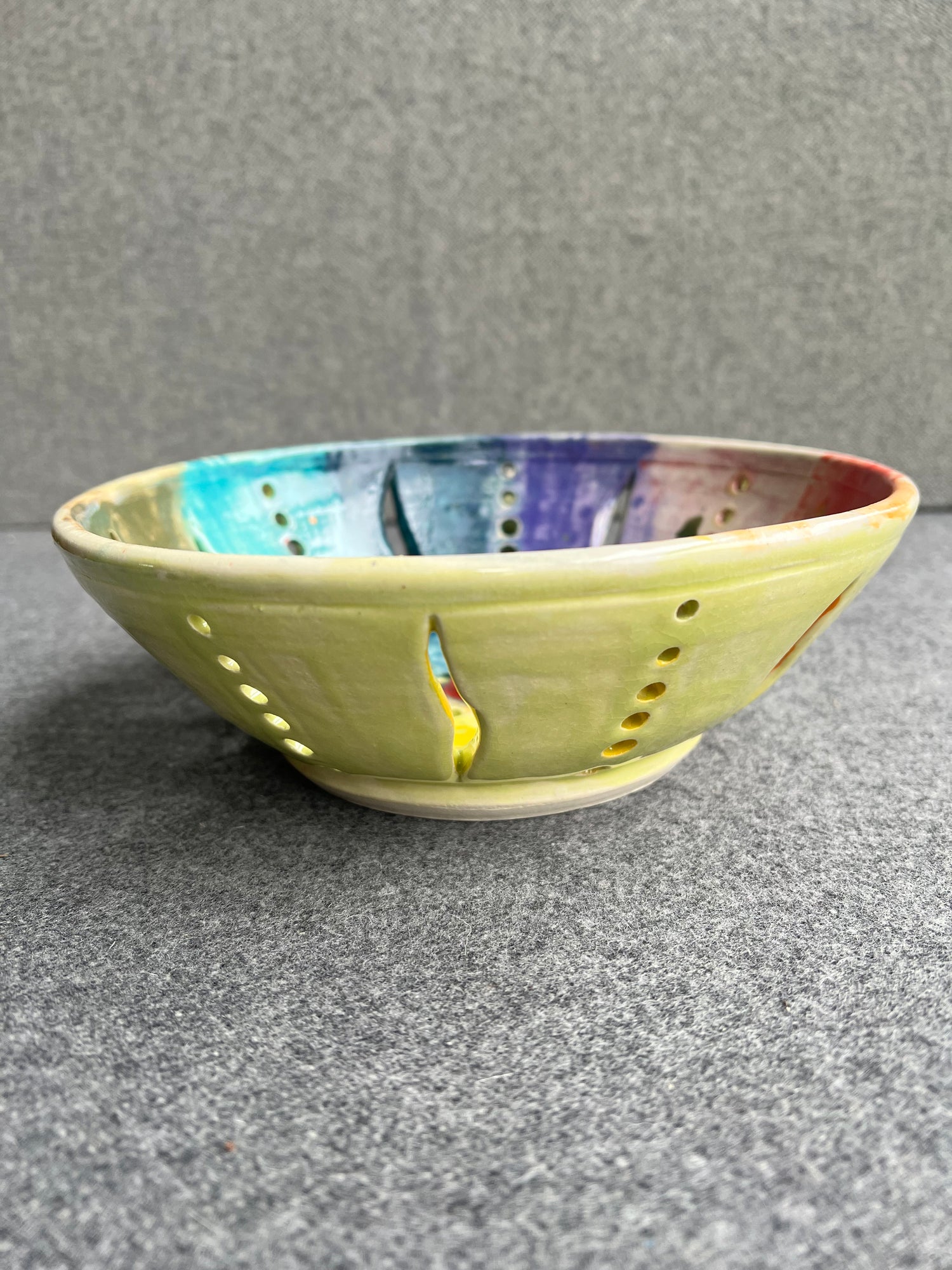 A side view of an intricately pierced bowl with a color wheel effect rainbow pattern and a small red heart in the center of the bowl sits in front of a grey background. The outside of the bowl is chartreuse green. 