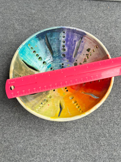 A bright pink ruler measuring 8 inches sits on top of an intricately pierced bowl with a color wheel effect rainbow pattern and a small red heart in the center of the bowl sits in front of a grey background. 