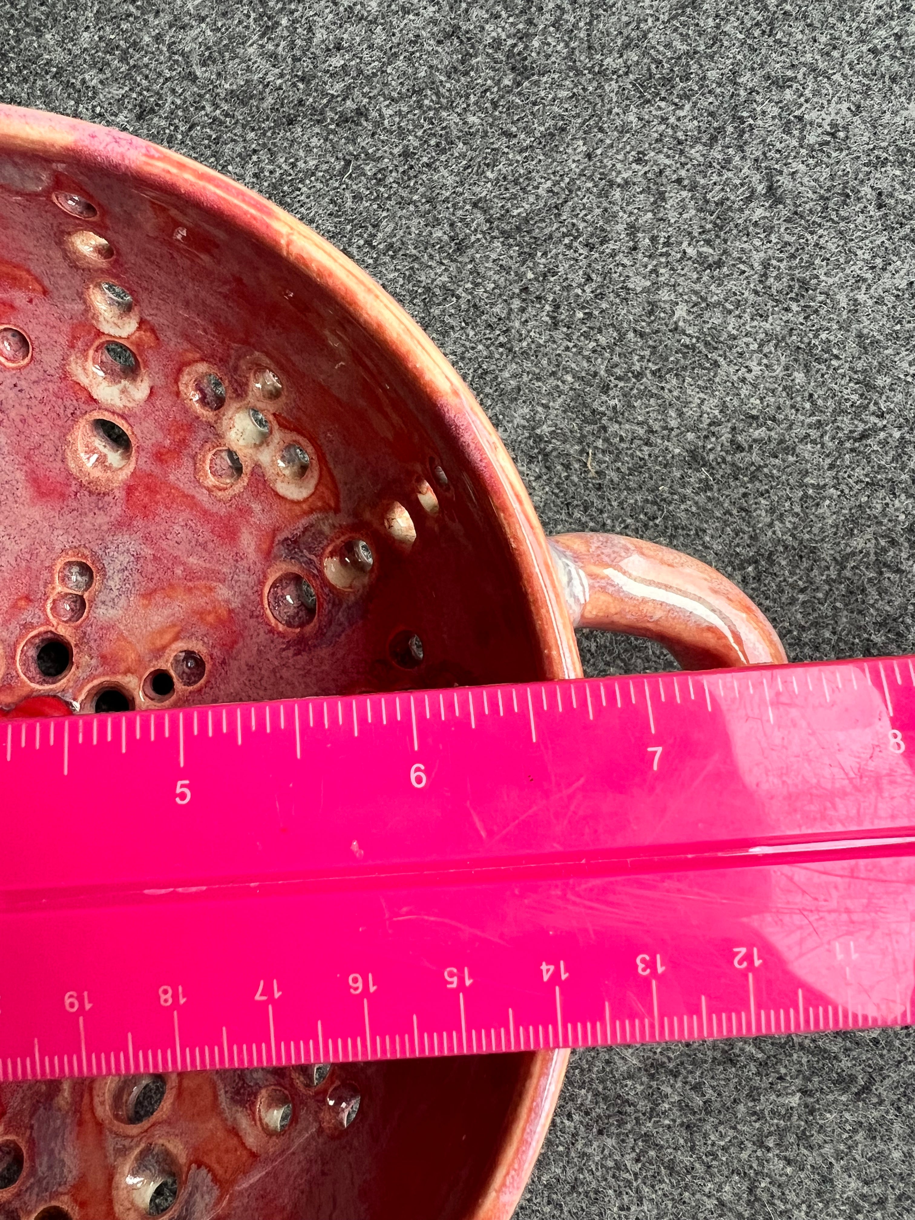 A bright pink ruler sits on top of a pink bowl with two small loop handles, and intricate pierced holes and a bright red heart at the base sits in front of a grey background. The ruler measure 6.75 inches.