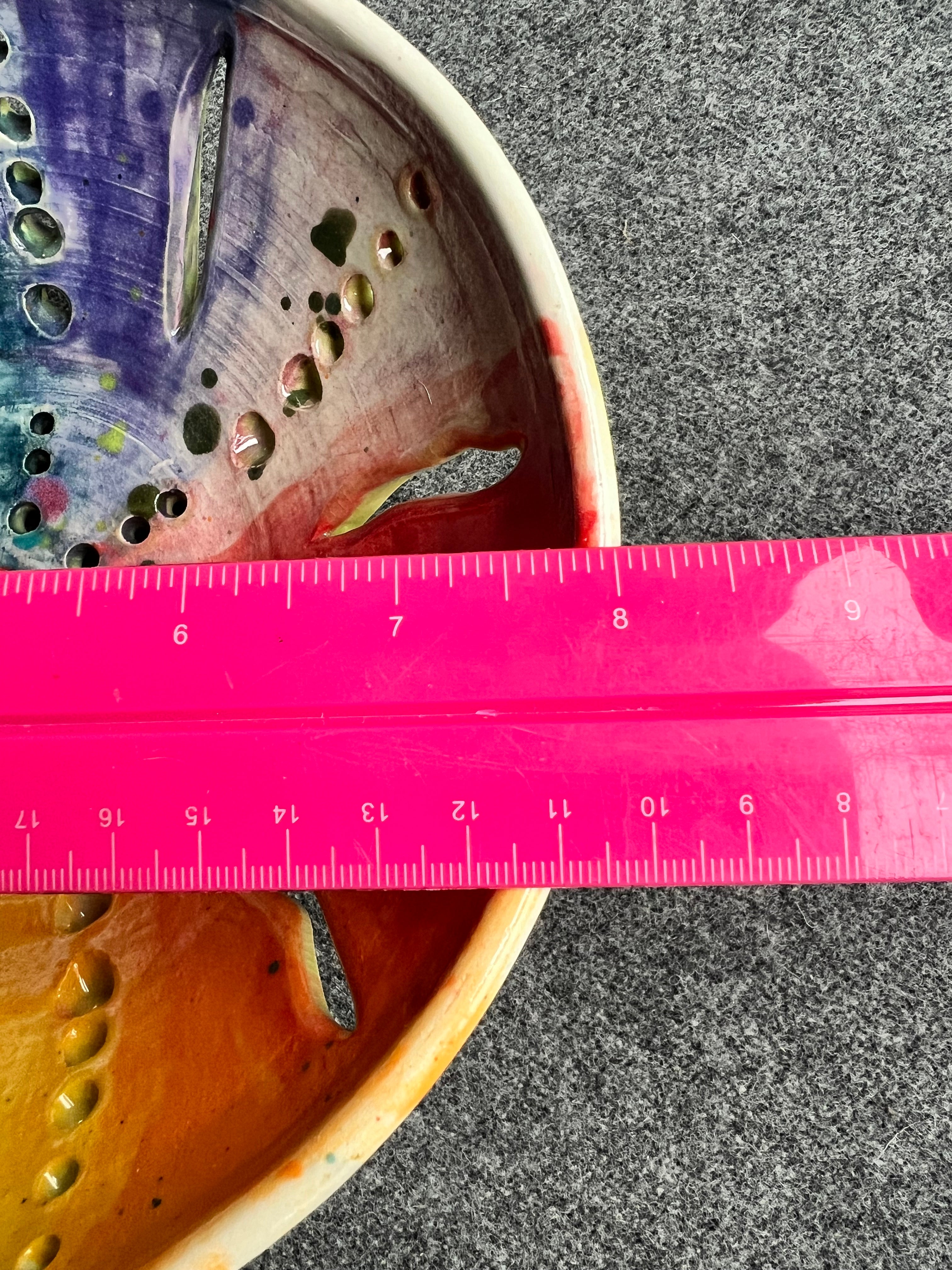 A close up of a bright pink ruler measuring 8 inches sits on top of an intricately pierced bowl with a color wheel effect rainbow pattern and a small red heart in the center of the bowl sits in front of a grey background. 