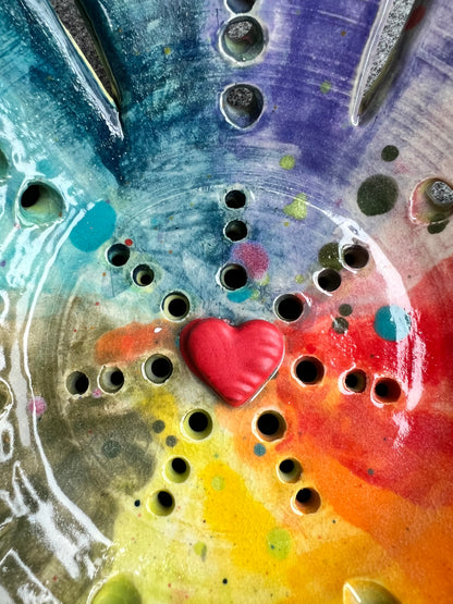 A close up of the center of an intricately pierced bowl with a color wheel effect rainbow pattern and a small red heart in the center of the bowl sits in front of a grey background. 