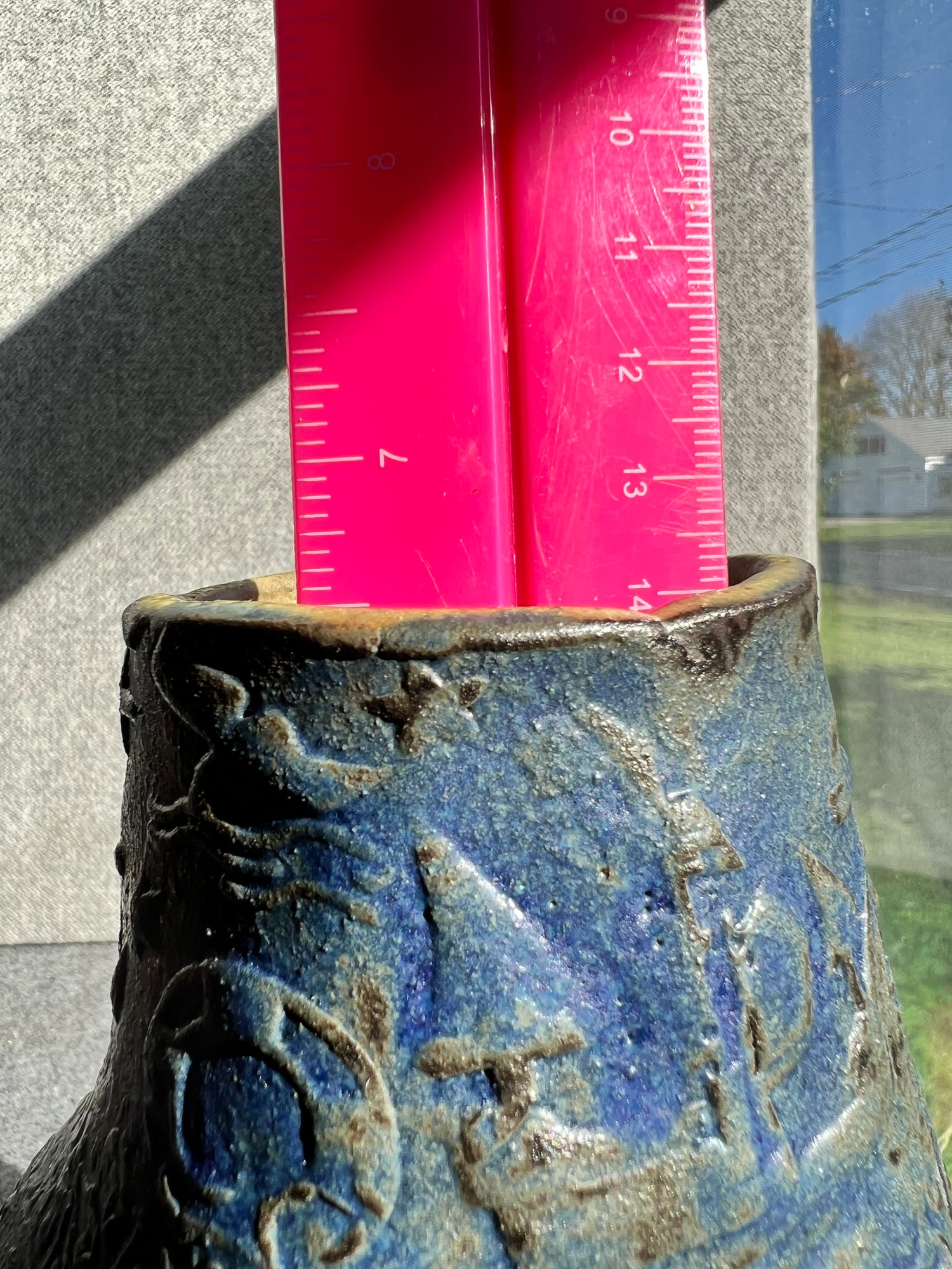 a three sided, handbill, green, brown, and blue ocean themed vase with a hot pink ruler inside of it measures 6.5 inches, and sits in front of a grey background. 