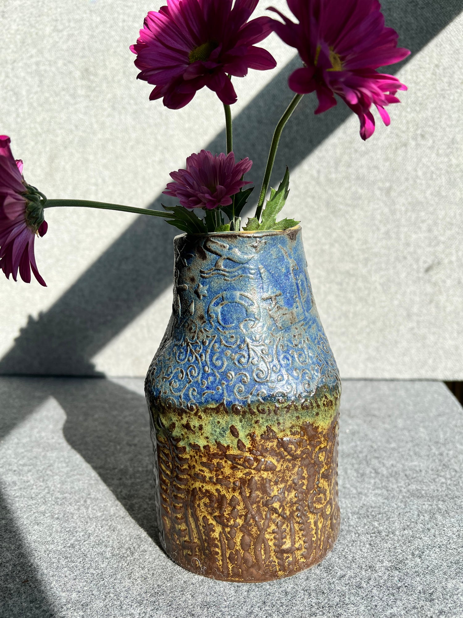 a three sided, handbill, green, brown, and blue ocean themed vase with four pink daisies sits in front of a grey background. 