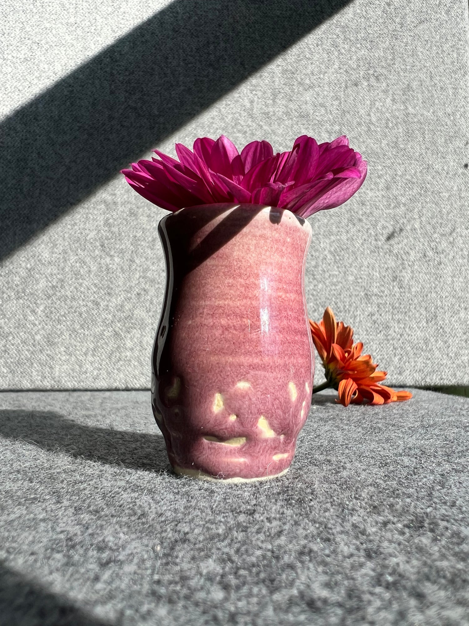 A curvaceous raspberry colored ceramic bud vase sits in front of an out of focus and a grey background. The glaze has crawled away from the surface of the vase leaving exposed areas of fired clay, leaving a mottled effect. 