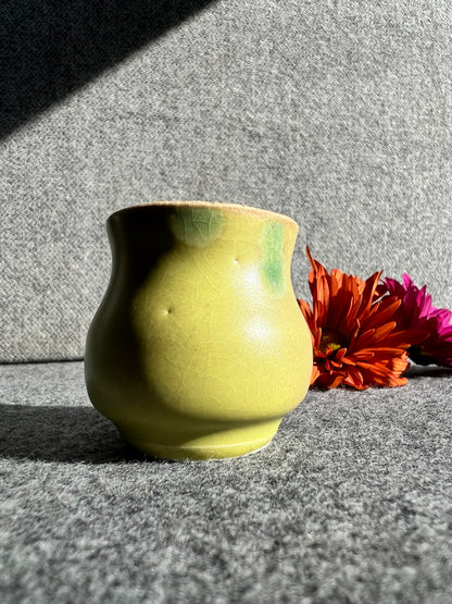A curvaceous, firefly yellow bud vase sits in front of two out of focus flowers and a grey background. 