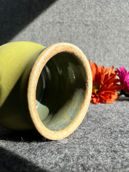 A curvaceous, firefly yellow bud vase lays on its side in front of two out of focus flowers and a grey background.