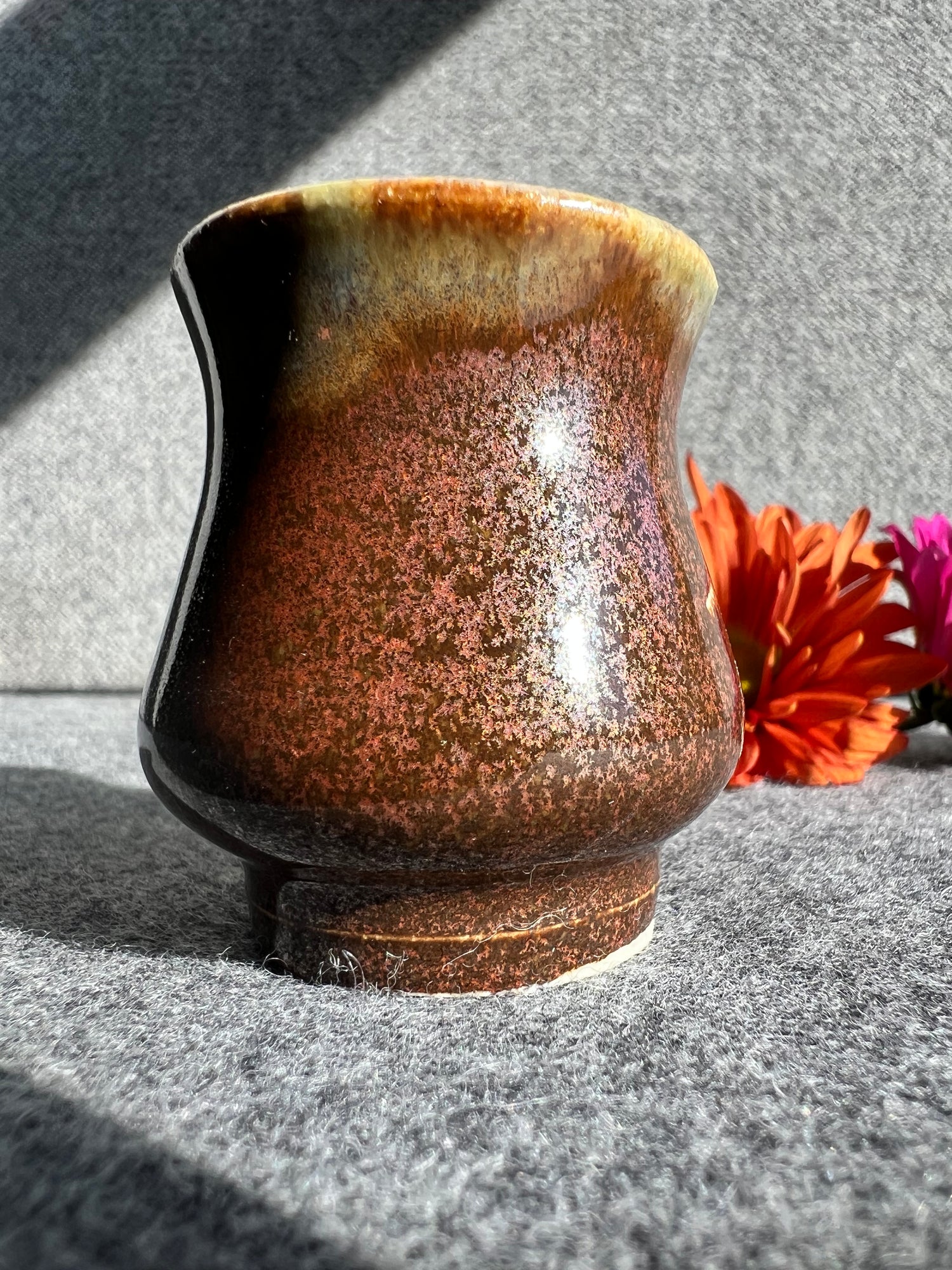 A curvaceous bud vase with a crystalline flecked copper-colored glaze sits in front of two out of focus flowers and a grey background. 