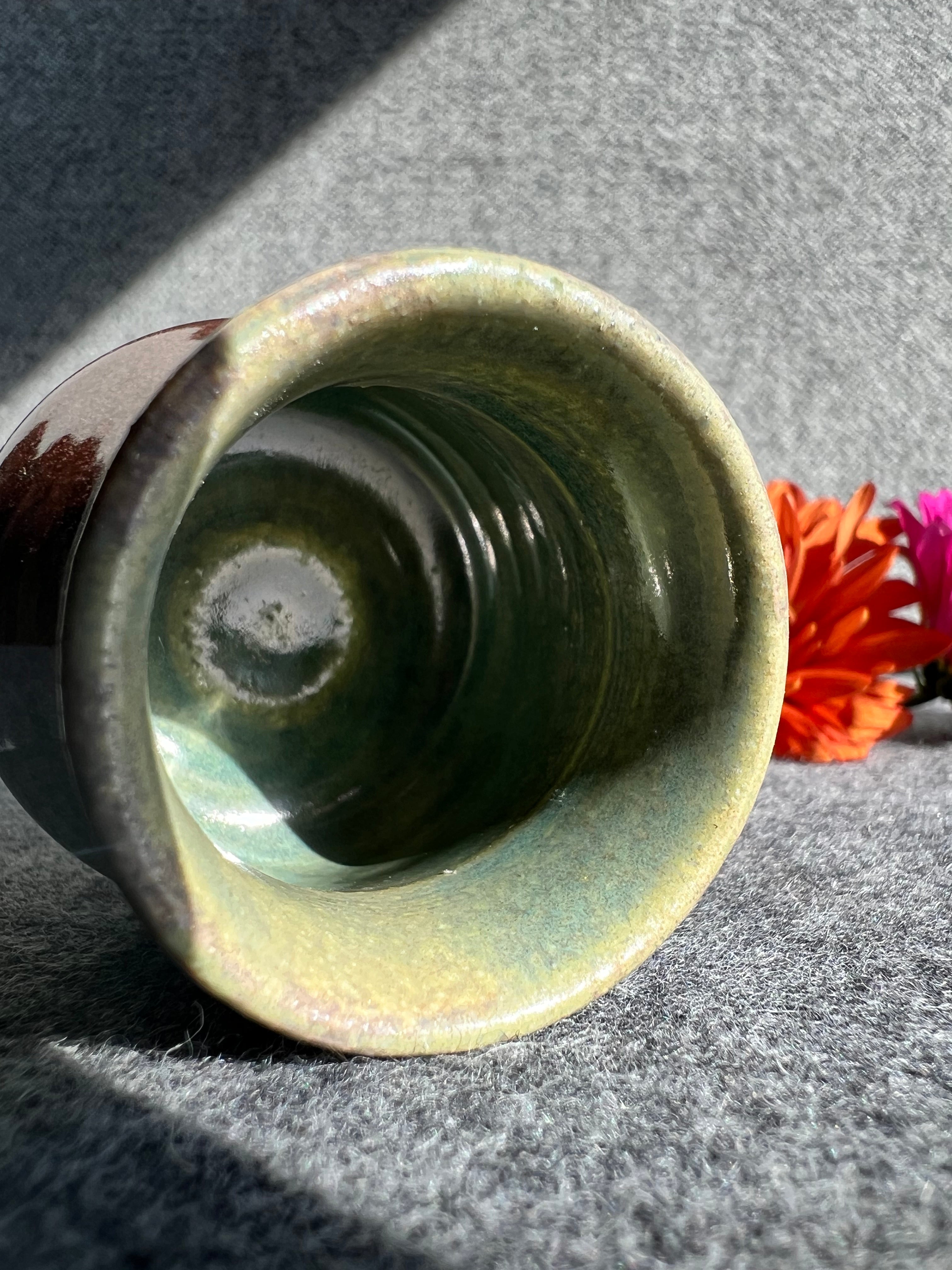 A curvaceous bud vase with a crystalline flecked copper-colored glaze lays on its side in front of two out of focus flowers and a grey background. 
