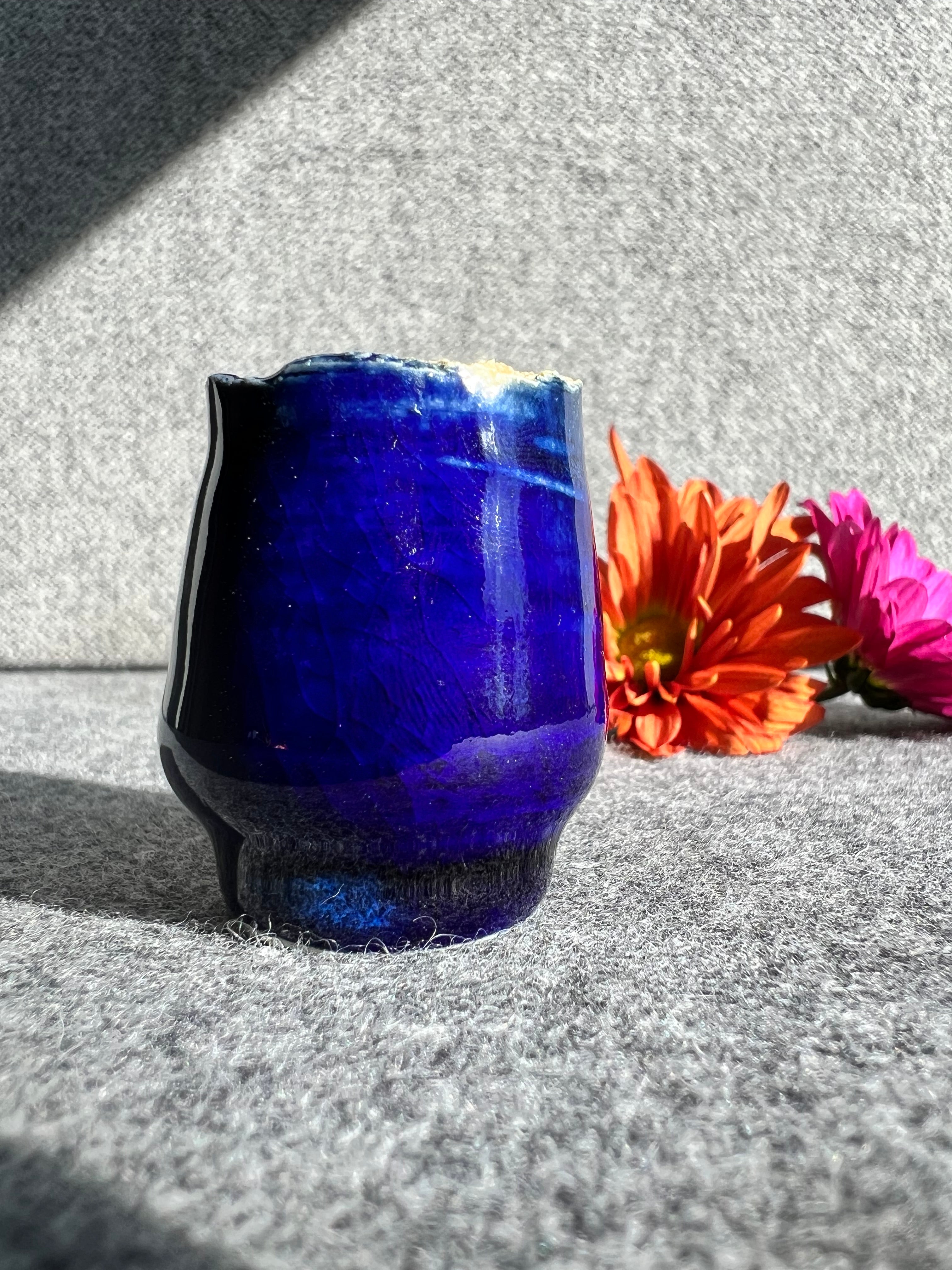 A cobalt blue bud vase with a textured rim sits in front of two out of focus flowers and a grey background. 
