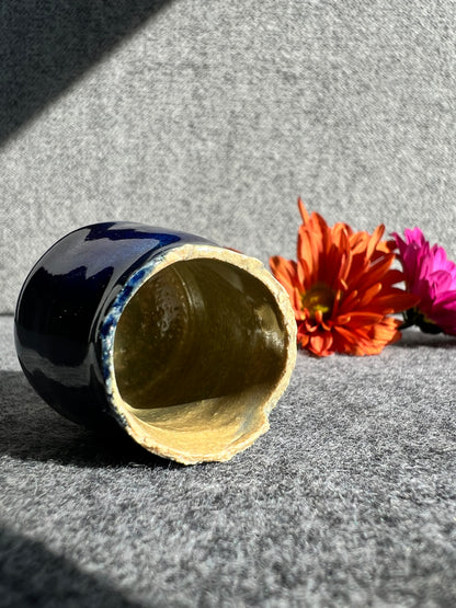 A cobalt blue bud vase with a textured rim lays on its side in front of two out of focus flowers and a grey background. 