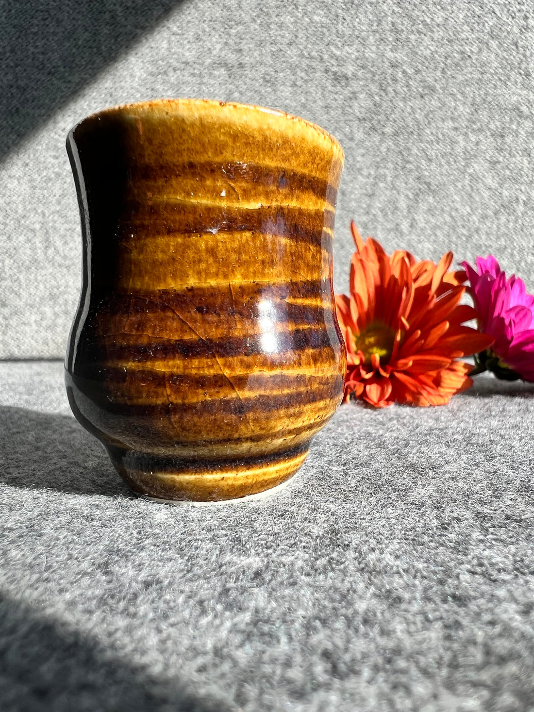 A rusty iron colored, glazed bud vase with a swirly texture sits in front of two out of focus flowers and a grey background. 