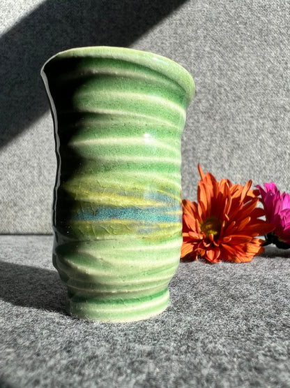A tall thin bud vase with a swirly texture and green and blue glaze sits in front of two out of focus flowers and a grey background. 