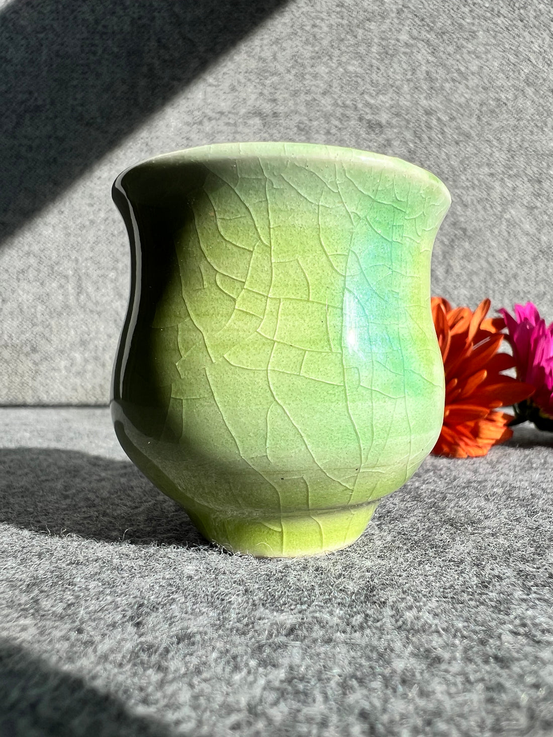 A bright green crackle glaze with hints of pale blue on a curvaceous bud vase sits in front of two out of focus flowers and a grey background. 