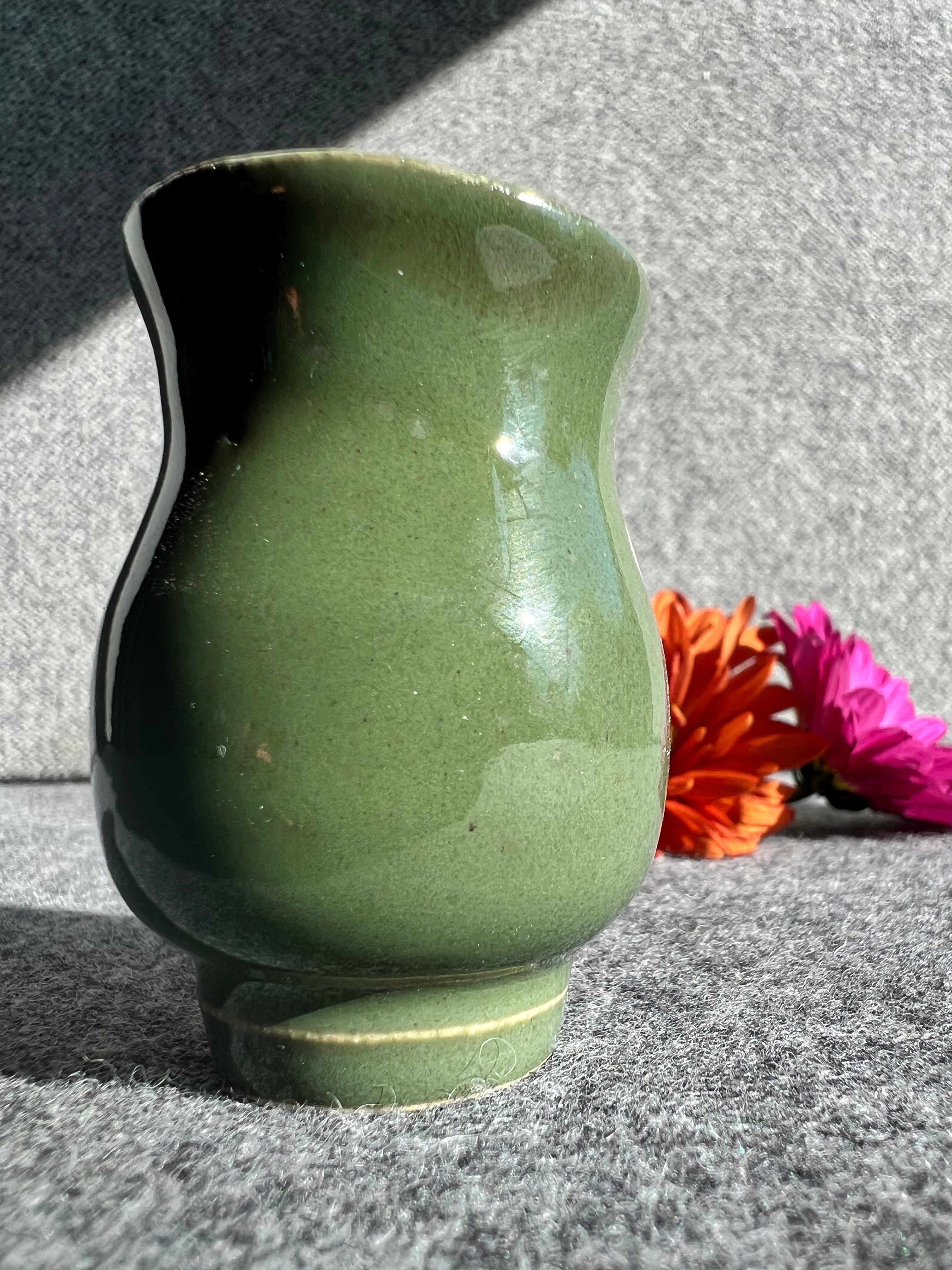A curvaceous, emerald green bud vase stands in front of two out of focus flowers and a grey background. 