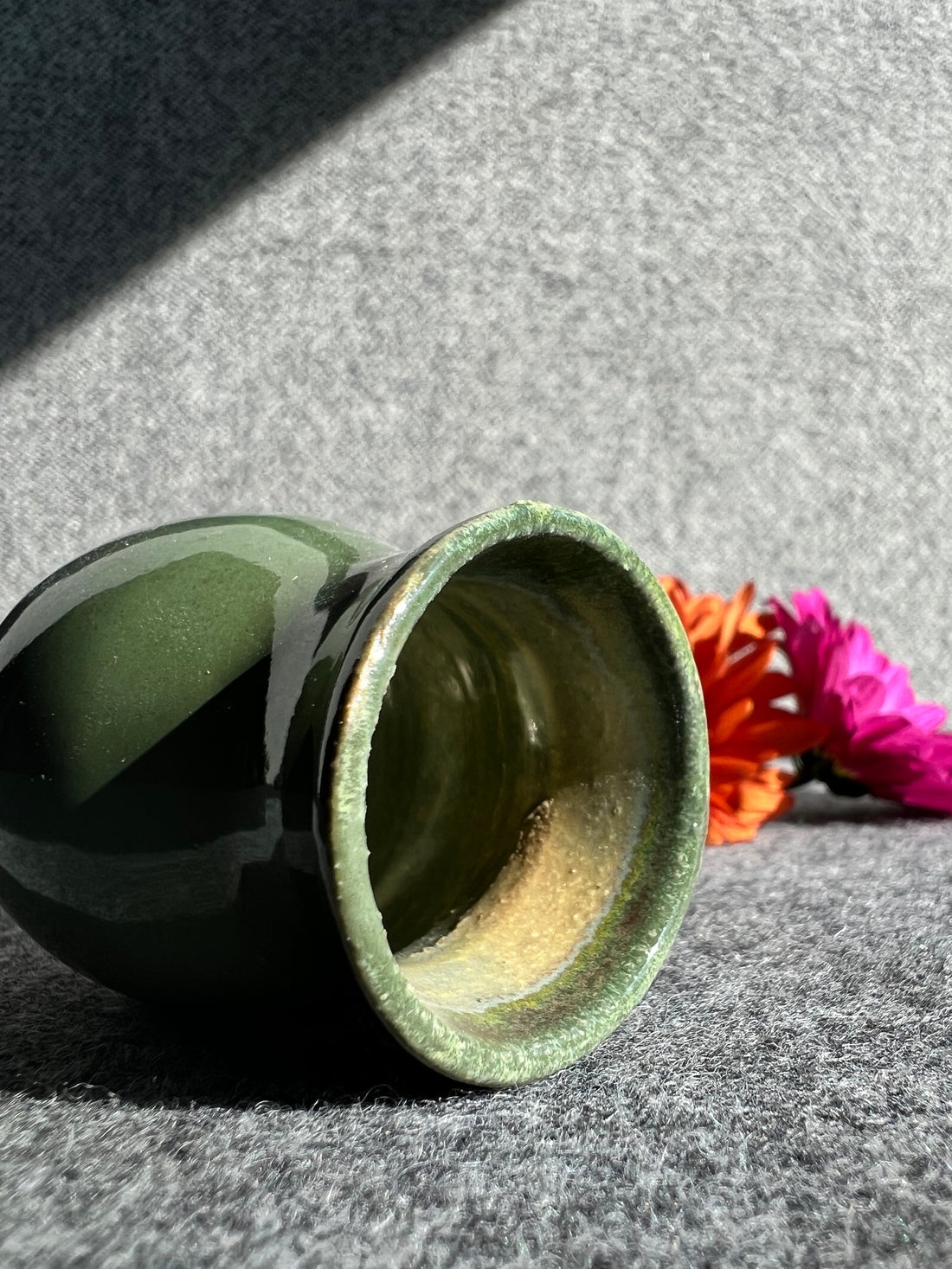 A curvaceous, emerald green bud vase lays on its side in front of two out of focus flowers and a grey background. 