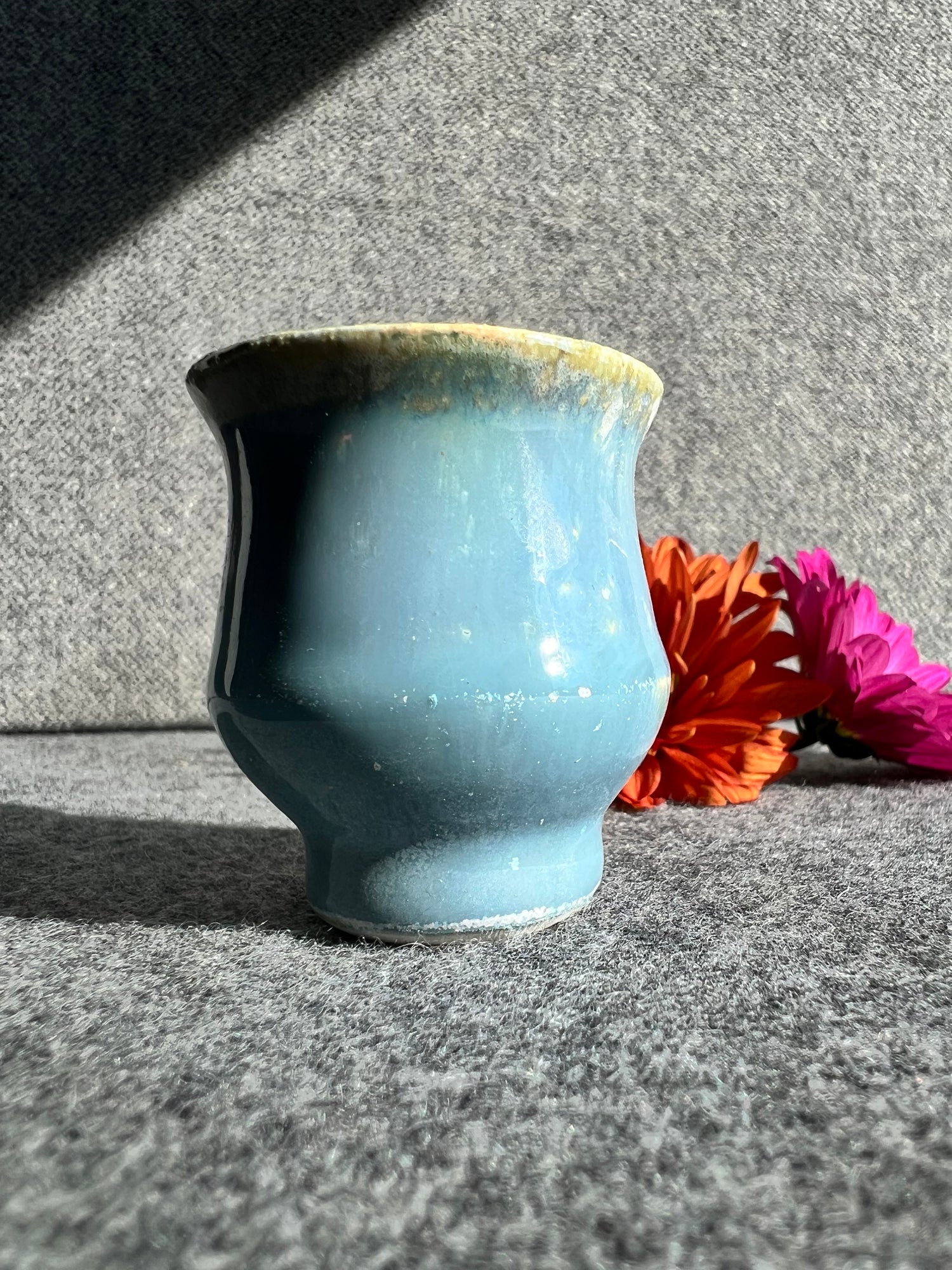 A curvaceous, robin egg blue bud vase stand in front of two out of focus flowers and a grey background. 