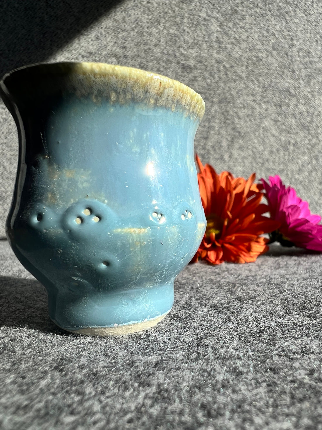 A curvaceous, robin egg blue bud vase with slight circular glaze defects stands in front of two out of focus flowers and a grey background. 