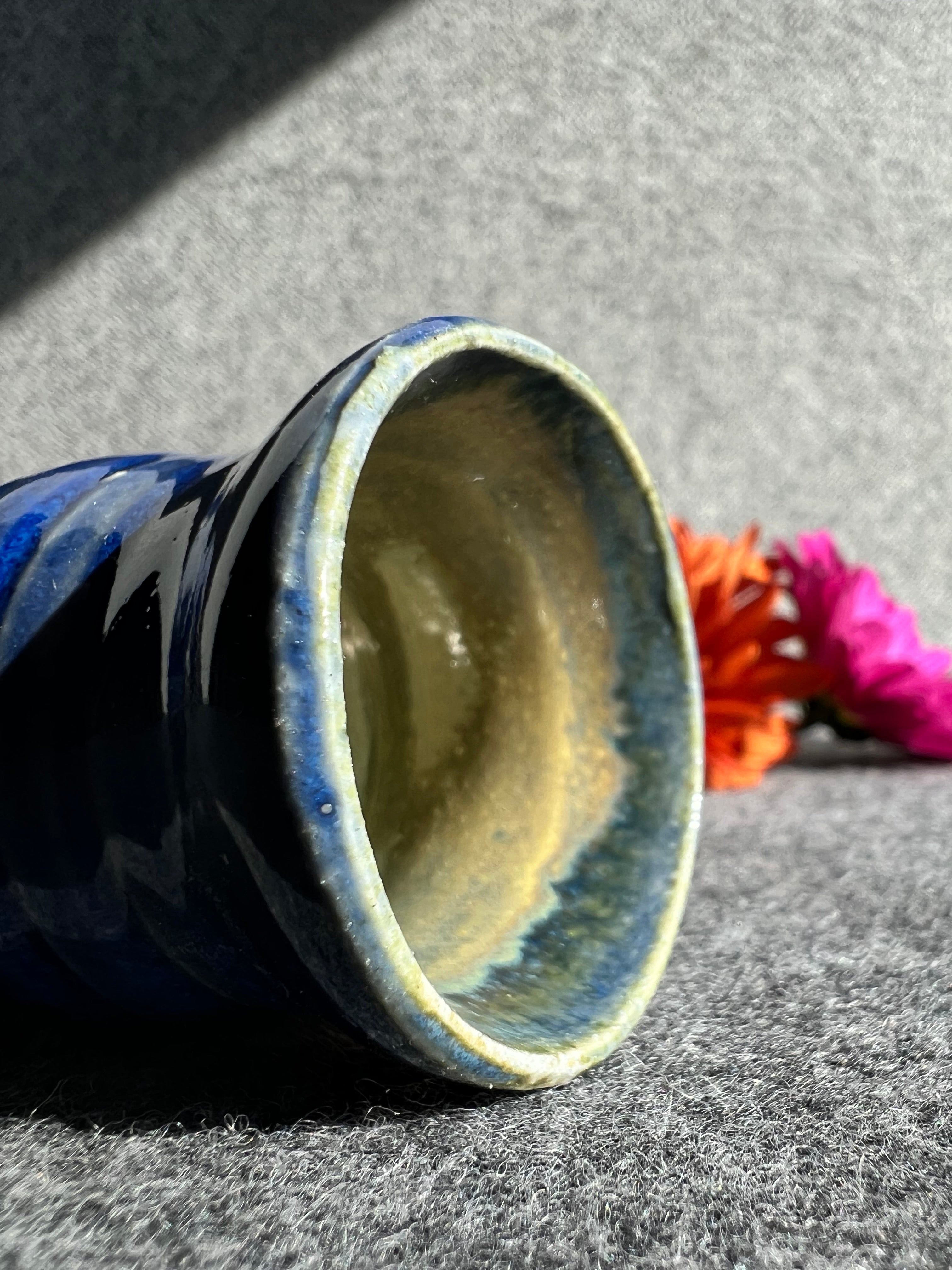 A bright blue bud vase lays on its side infant of a grey background with two out of focus flowers in the distance. 