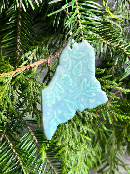 A blue, state of Maine ornament with a leafy textured surface and a clear crackle glaze hangs in front of cedar and balsam branches. 