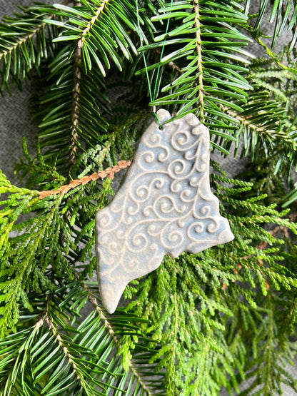 A blue, state of Maine ornament with a curly, swirly textured surface and a clear crackle glaze hangs in front of cedar and balsam branches. 