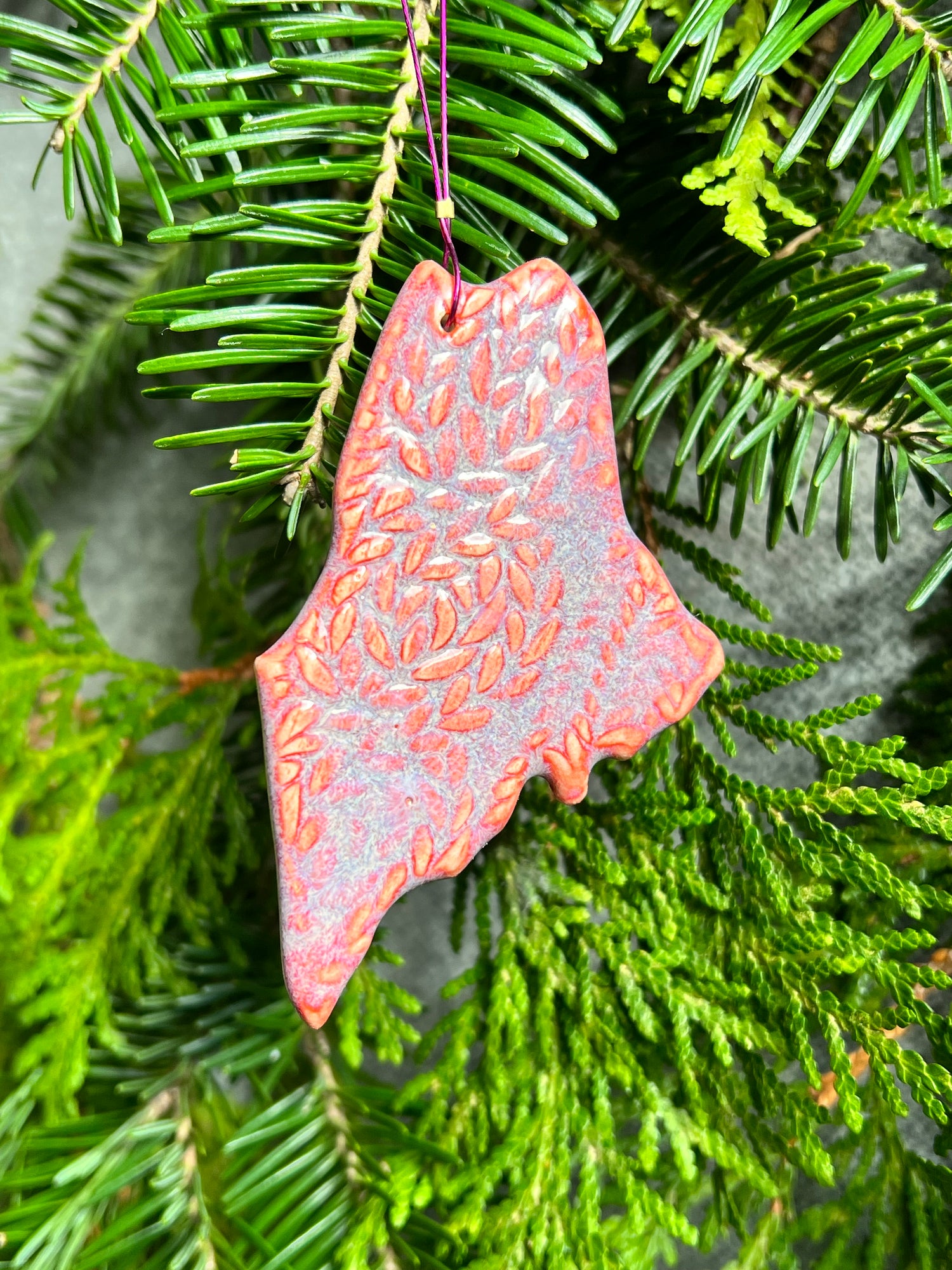 A pink and lavender, state of Maine ornament with a cabled knit textured surface and a clear crackle glaze hangs in front of cedar and balsam branches. 