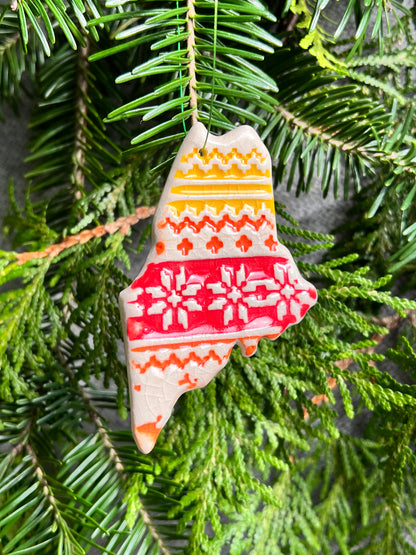 A red, orange, and yellow state of Maine ornament with a nordic textured surface and a clear crackle glaze hangs in front of cedar and balsam branches. 