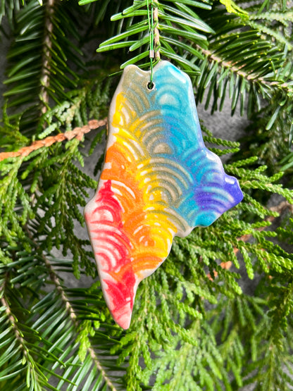 A state of Maine ornament with a rainbow of painted vertical stripes and a rainbow textured surface and a clear crackle glaze hangs in front of cedar and balsam branches. 