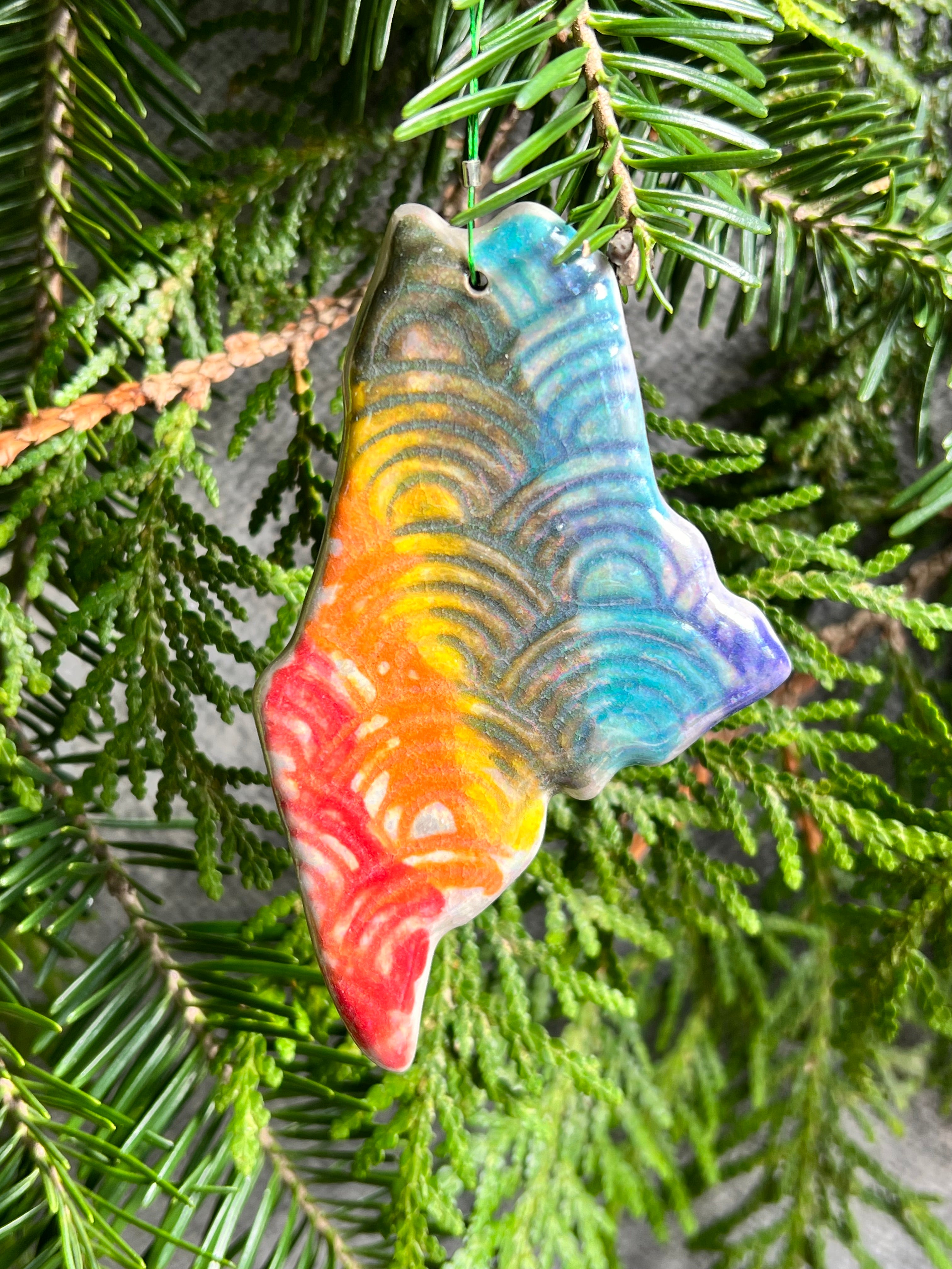 A state of Maine ornament with a rainbow of painted vertical stripes and a rainbow textured surface and a clear crackle glaze hangs in front of cedar and balsam branches.