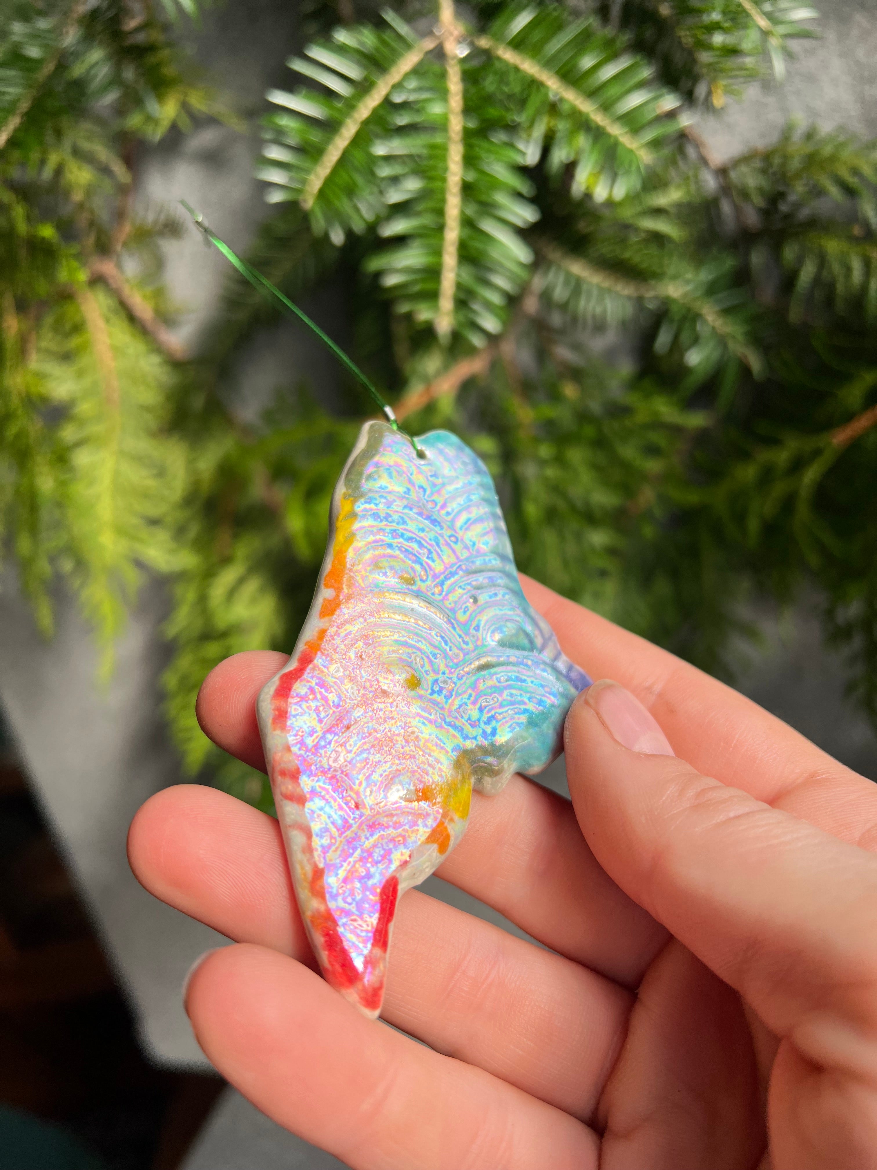 A state of Maine ornament with a rainbow of painted vertical stripes and a rainbow textured surface is held at an angle in front of cedar and balsam branches to show off the rainbow iridescent sheen on the surface.
