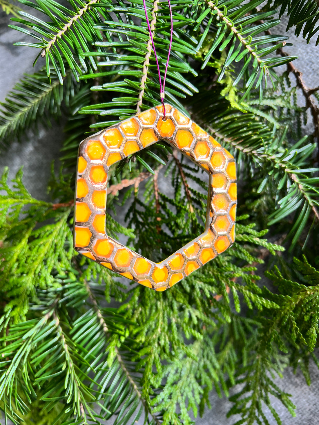 A yellow, orange, and gold open hexagon with a textured honeycomb surface ornament hangs in front of cedar and balsam branches.