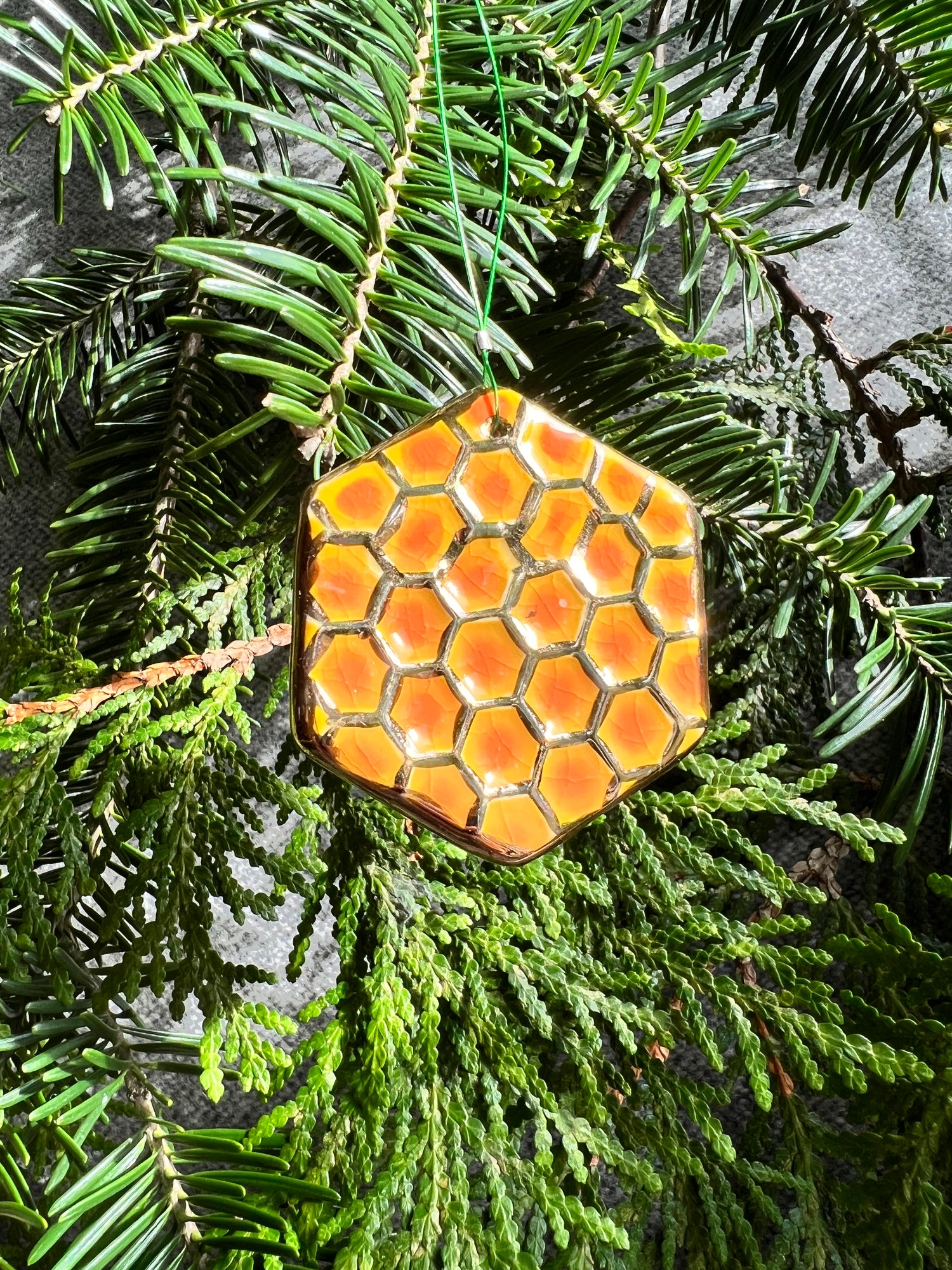 A yellow, orange, and gold hexagon with a textured honeycomb surface ornament hangs in front of cedar and balsam branches.